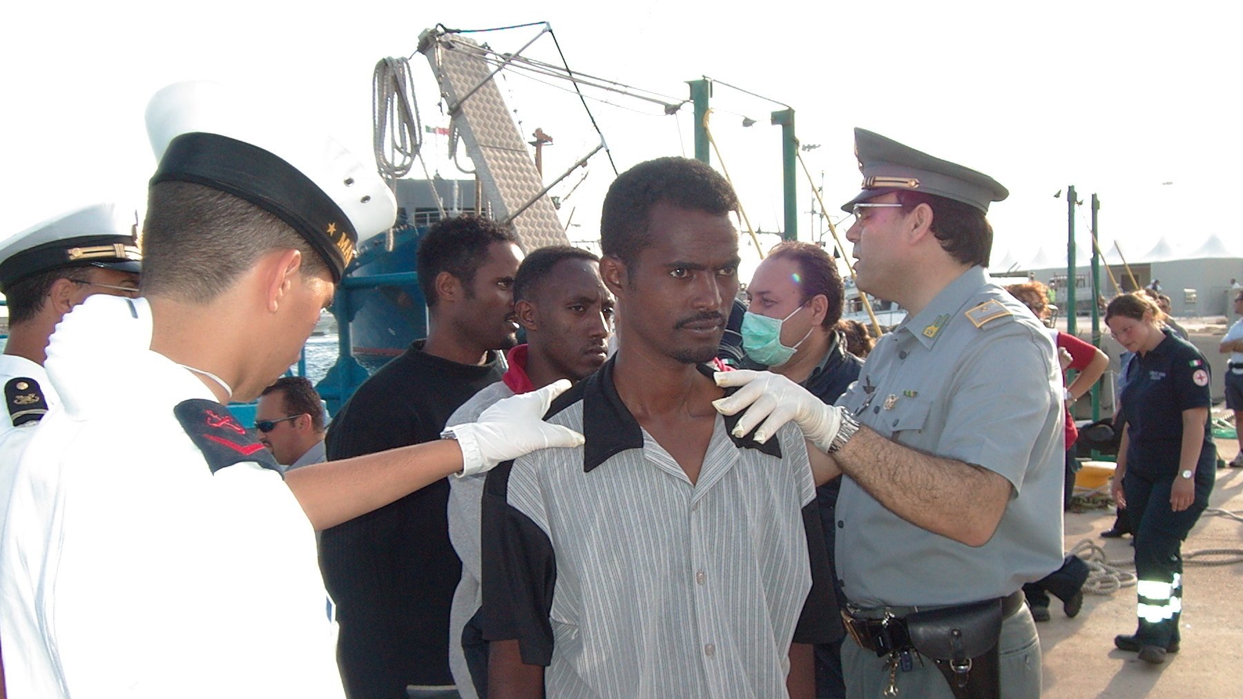 L'image montre une scène avec plusieurs personnes. Au premier plan, un homme se tient debout, apparemment en train d'être examiné par des agents portant des uniformes. Il y a un groupe d'autres personnes en arrière-plan, semblant également être supervisées. L'environnement suggère que cela se passe dans un port ou une zone maritime, avec des bateaux et des structures portuaires visibles. Les agents semblent s'occuper de la situation de manière attentive.