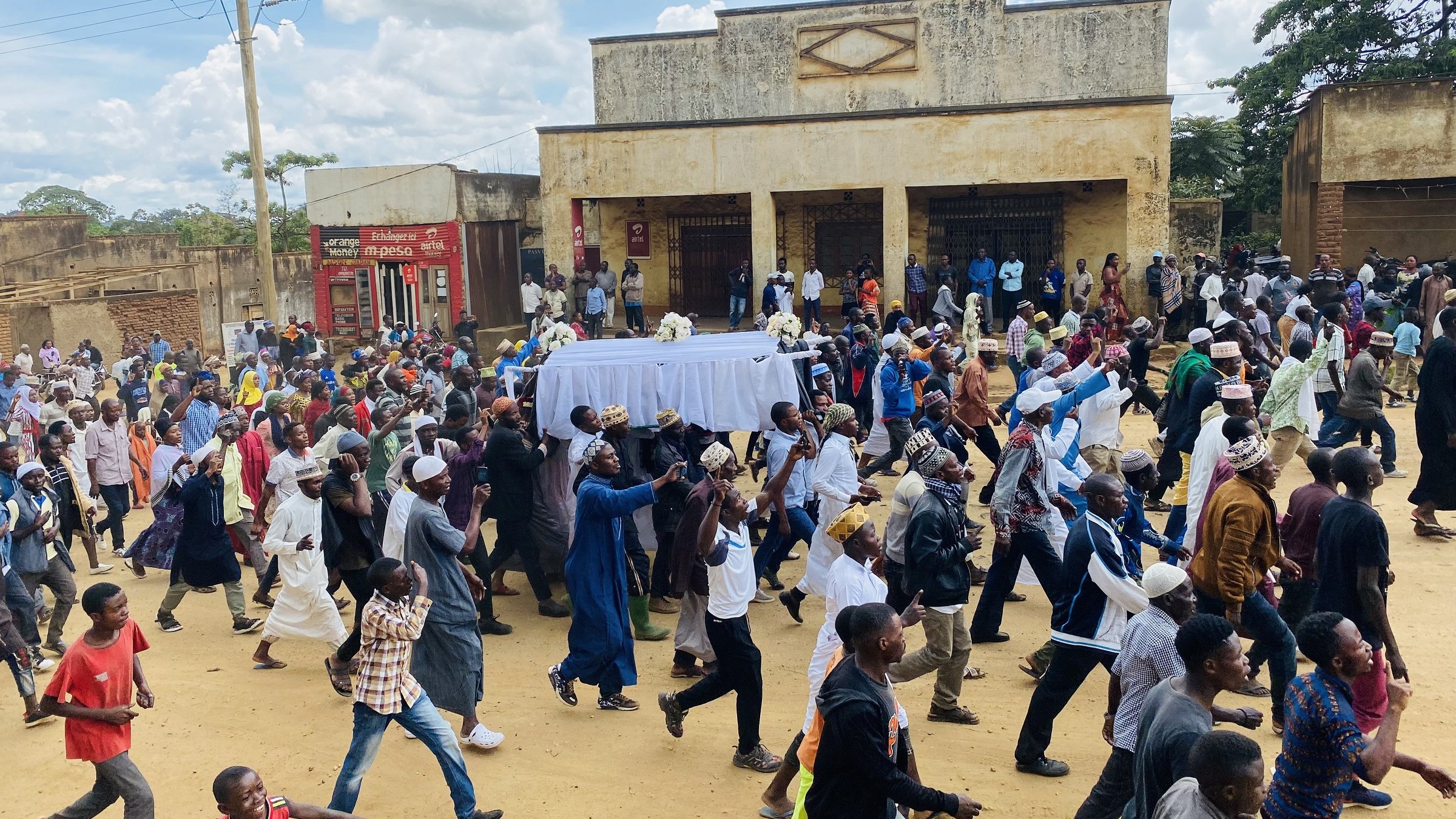 L'image montre une grande foule de personnes marchant dans une rue, probablement lors d'un événement ou d'une procession. Au centre, il y a un chariot couvert d'un drap blanc, et les gens semblent marcher avec une certaine ferveur. On peut apercevoir différents styles vestimentaires, suggérant une diversité parmi les participants. En arrière-plan, des bâtiments sont visibles, ainsi qu'un ciel partiellement nuageux. L'atmosphère semble collective et déterminée.