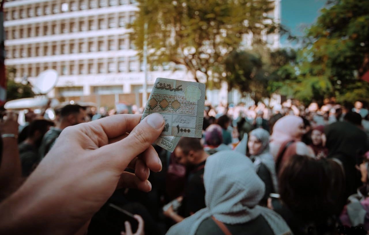 L'image montre une main tenant un billet de banque marocain au premier plan, tandis qu'à l'arrière-plan, une foule est rassemblée. On aperçoit des personnes aux tenues variées, certaines portant des foulards. Le cadre semble être un événement public, probablement une manifestation ou une célébration, avec des bâtiments modernes en arrière-plan et des arbres qui ajoutent une touche de verdure à la scène. L'atmosphère semble dynamique et chargée d'énergie collective.