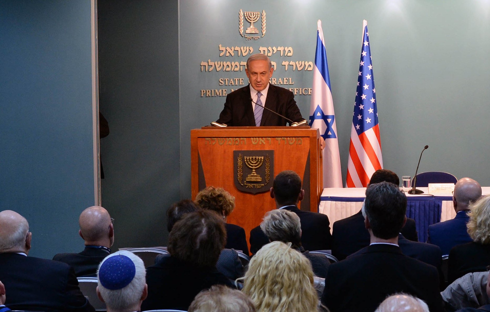 L'image montre un homme en train de s'adresser à un public depuis un podium marqué du sceau d'Israël et des États-Unis. Il y a des personnes assises derrière lui, écoutant son discours. On peut voir des drapeaux israélien et américain en arrière-plan. L'ambiance semble formelle, probablement dans le cadre d'une conférence ou d'une réunion officielle.