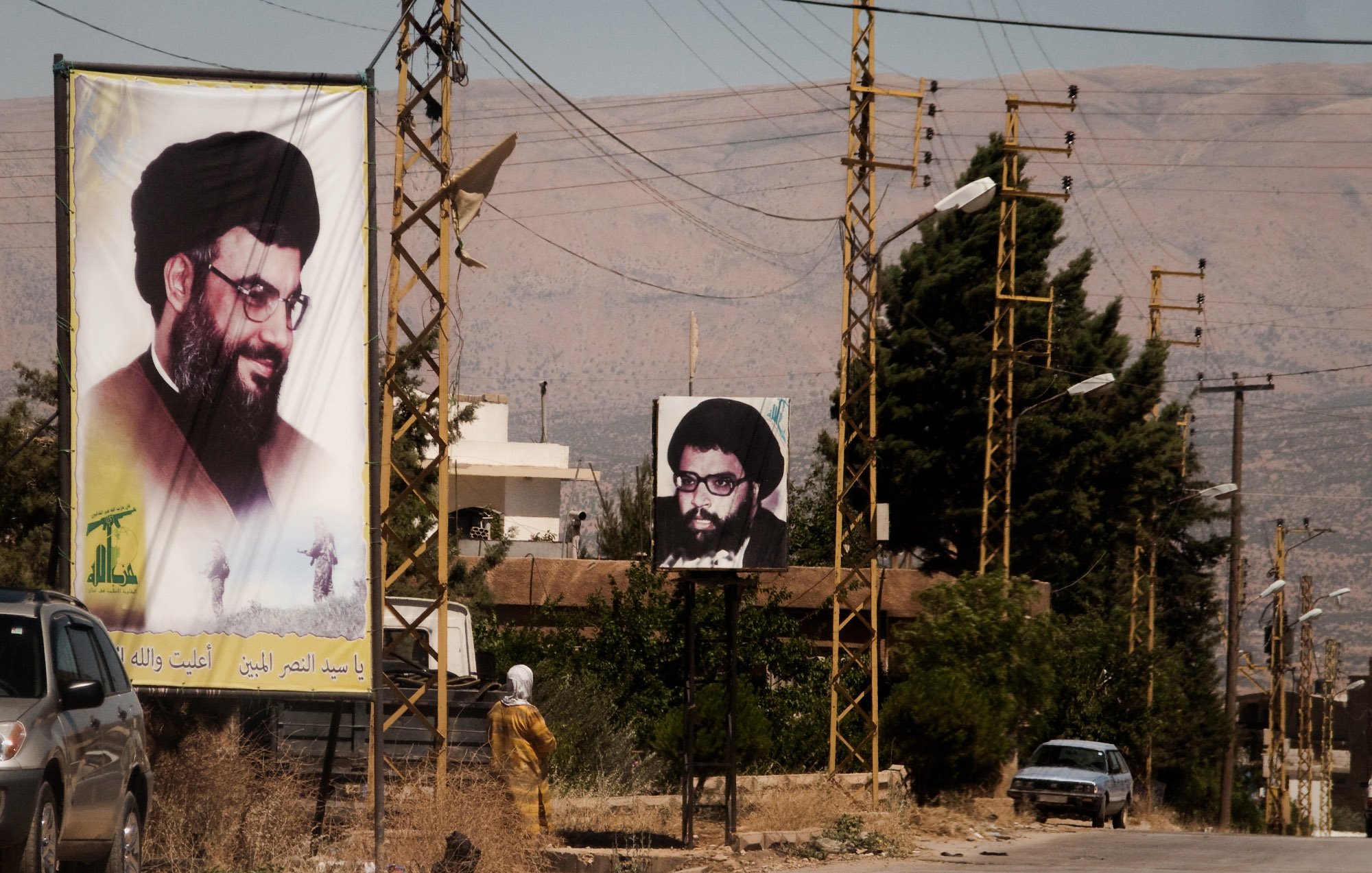 L'image montre une rue avec des panneaux d'affichage sur lesquels figurent des portraits d'un homme portant des lunettes et une barbe, associé à une ambiance rurale. On peut également voir des lignes électriques et quelques bâtiments en arrière-plan. À gauche, un panneau est en couleur et à droite, un autre est en noir et blanc. La scène semble être située dans une région montagneuse. C'est une illustration d'un espace public où des affiches politiques ou religieuses peuvent être observées.