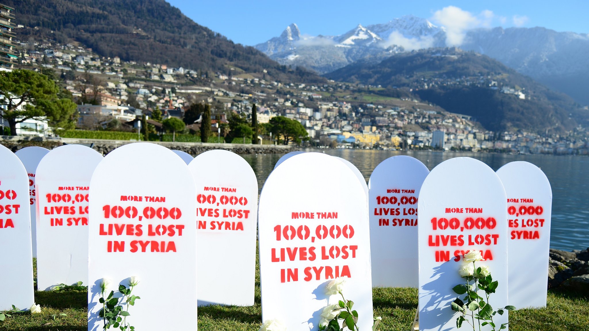 L'image montre un rassemblement commémoratif avec plusieurs pierres tombales blanches disposées sur un terrain vert. Chaque pierre tombale est marquée de rouge avec des inscriptions telles que "More than 100,000 lives lost in Syria". À côté des pierres tombales, il y a des roses ou des plantes, symbolisant probablement la mémoire des vies perdues. En arrière-plan, on peut voir une vue magnifique de montagnes et d'une ville au bord de l'eau. L'ambiance générale semble être solennelle et réfléchissante, soulignant une tragédie humanitaire.