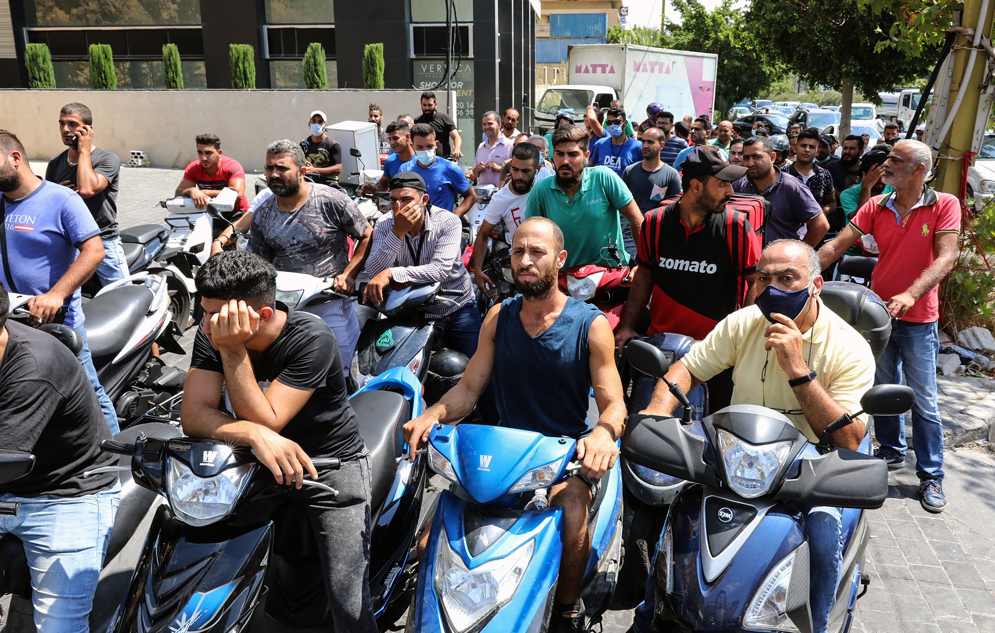 L'image montre un groupe de personnes, principalement des hommes, rassemblés sur des motos dans une file d'attente. Ils semblent attendre pour quelque chose, peut-être un service ou un approvisionnement. Certains affichent des expressions de frustration ou d'impatience. En arrière-plan, on peut voir des bâtiments et des arbres qui ajoutent au cadre urbain. L'atmosphère donne une impression de tension liée à une situation d'attente prolongée.
