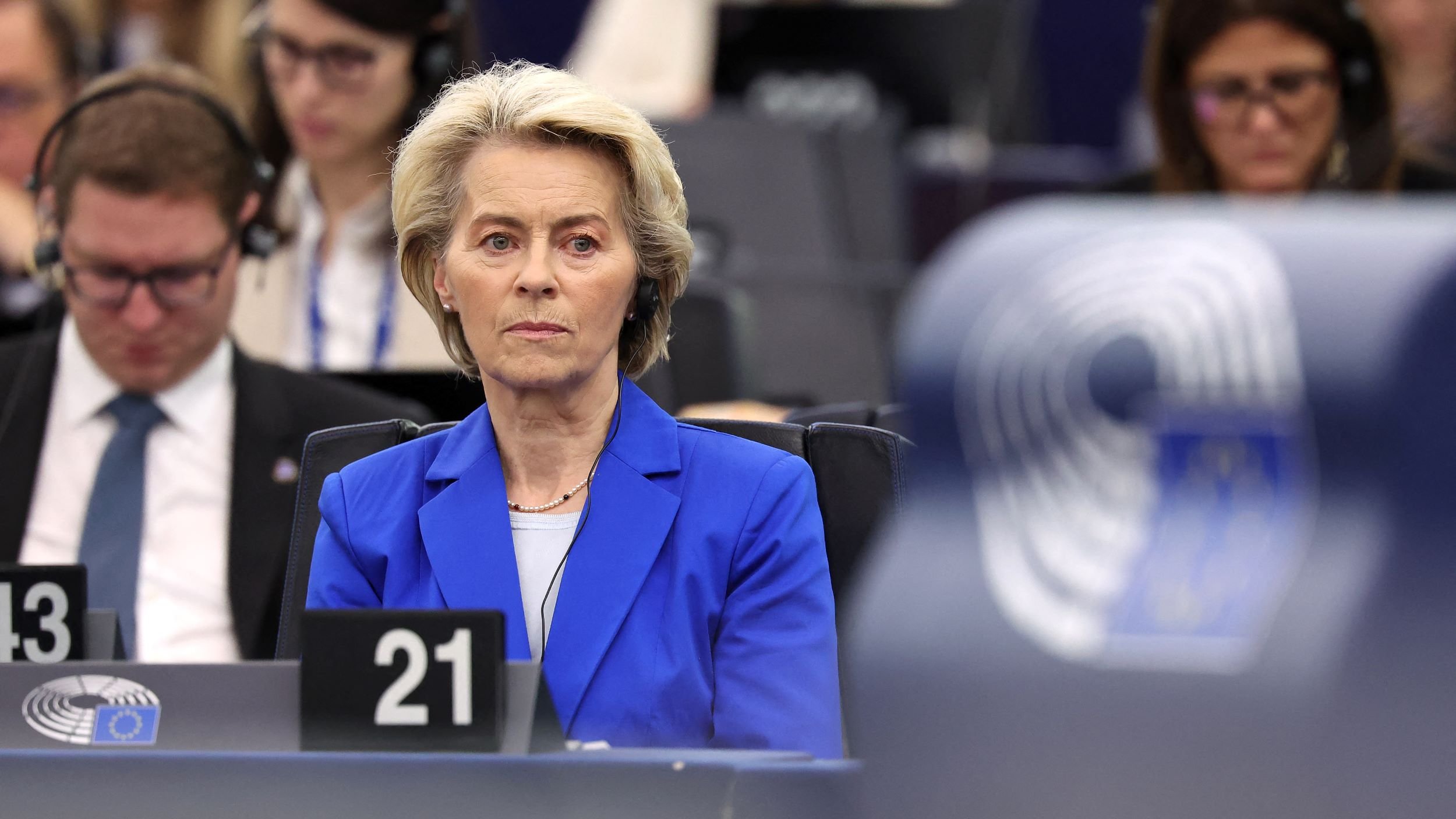 L'image montre une femme assise dans un environnement de réunion, probablement au sein d'une institution européenne, comme le Parlement européen. Elle porte un blazer bleu et semble concentrée sur un sujet abordé lors de la séance. En arrière-plan, on aperçoit d'autres personnes, également attentives. L'atmosphère paraît formelle et sérieuse, et des équipements audio sont visibles, suggérant une translation ou un écoute attentive des débats.
