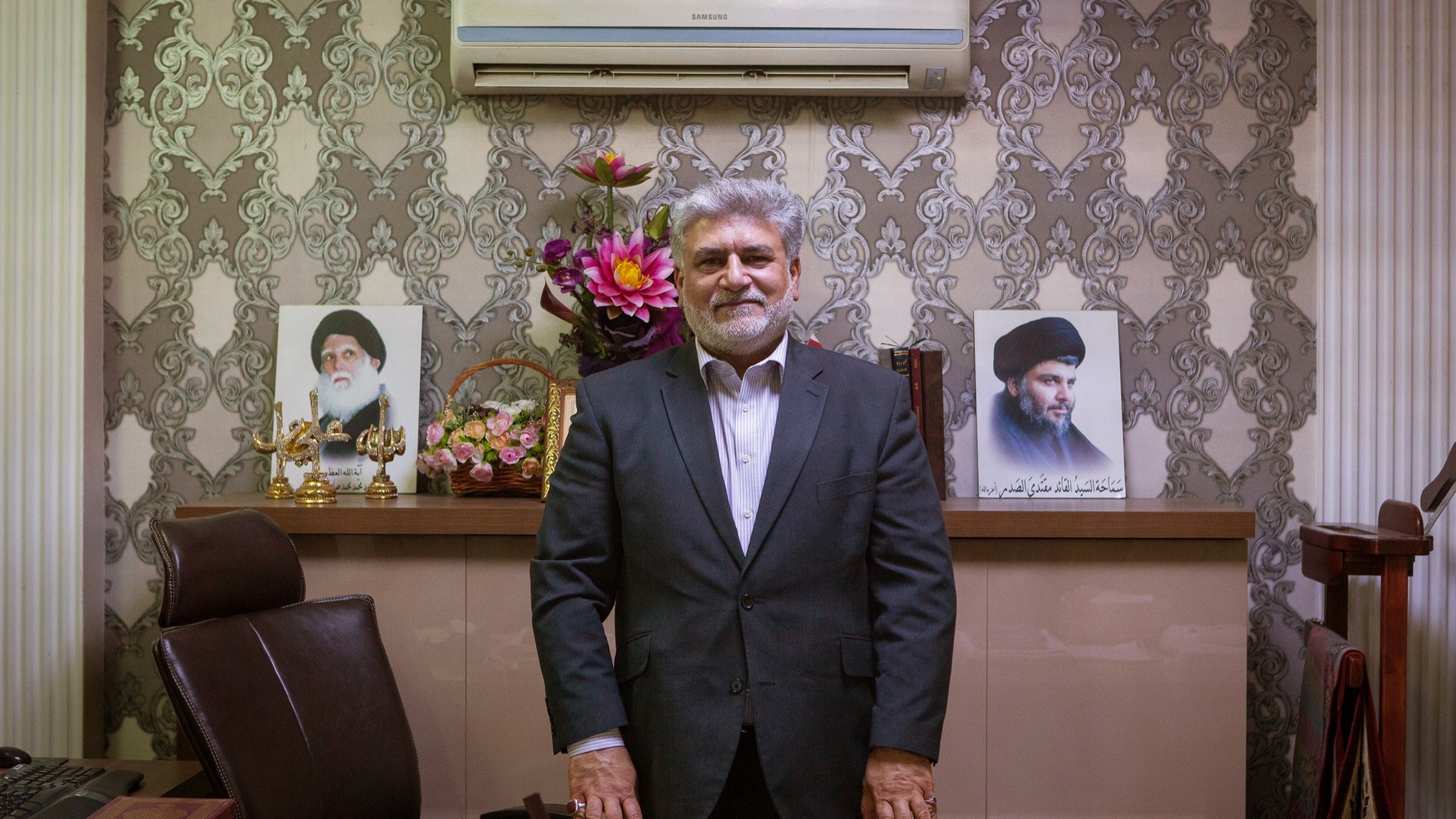 L'image montre un homme debout devant un bureau bien aménagé. Il porte un costume sombre et un sourire. Derrière lui, on peut voir des portraits de deux figures, ainsi qu'un arrangement floral. Le décor est élégant avec des murs ornés de motifs. L'ensemble dégage une atmosphère de professionnalisme et d'autorité.