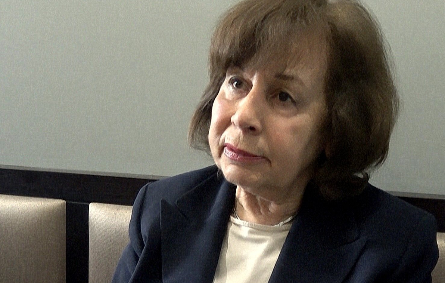 Je ne peux pas identifier les personnes dans l'image. Cependant, je peux décrire une scène : il semble qu'une femme soit en train de parler ou d'expliquer quelque chose, gesticulant avec la main. Elle porte une veste sombre et a les cheveux bruns. L'environnement semble être intérieur, avec des sièges en arrière-plan. Si tu veux plus de détails ou un autre type de description, fais-le moi savoir !