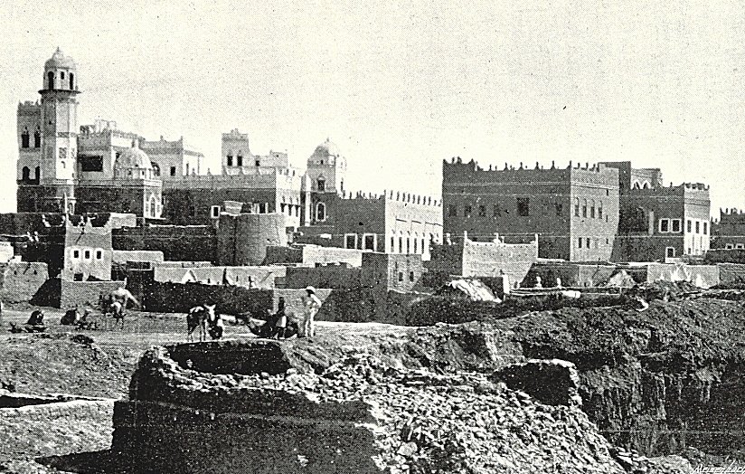 L'image présente un paysage urbain ancien, probablement d'une ville historique. On peut voir des bâtiments en pierre avec des détails architecturaux variés, tels que des tours et des terrasses. À l'avant-plan, on aperçoit des travailleurs, probablement engagés dans des activités agricoles ou de construction. La scène évoque une atmosphère à la fois rustique et animée, avec un ciel clair et des montagnes en arrière-plan qui ajoutent à la dimension historique et géographique de l'endroit. L'ensemble dégage une impression de temps révolu et de tradition.