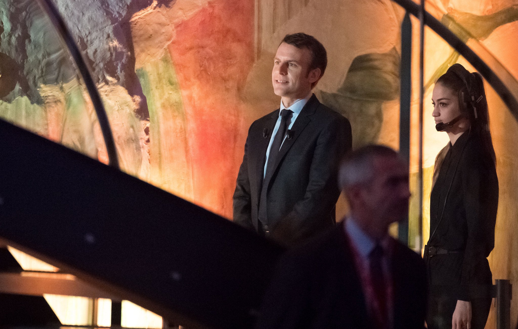 L'image montre un homme en costume, debout sur une scène, avec une femme à ses côtés qui porte un microphone et un ouïe. L'arrière-plan présente des couleurs et des formes abstraites, suggérant un environnement artistique ou un événement important. L'homme semble attentif, tandis que la femme semble concentrée sur la situation. Il y a également un autre homme en arrière-plan qui marche, suggérant une ambiance dynamique.