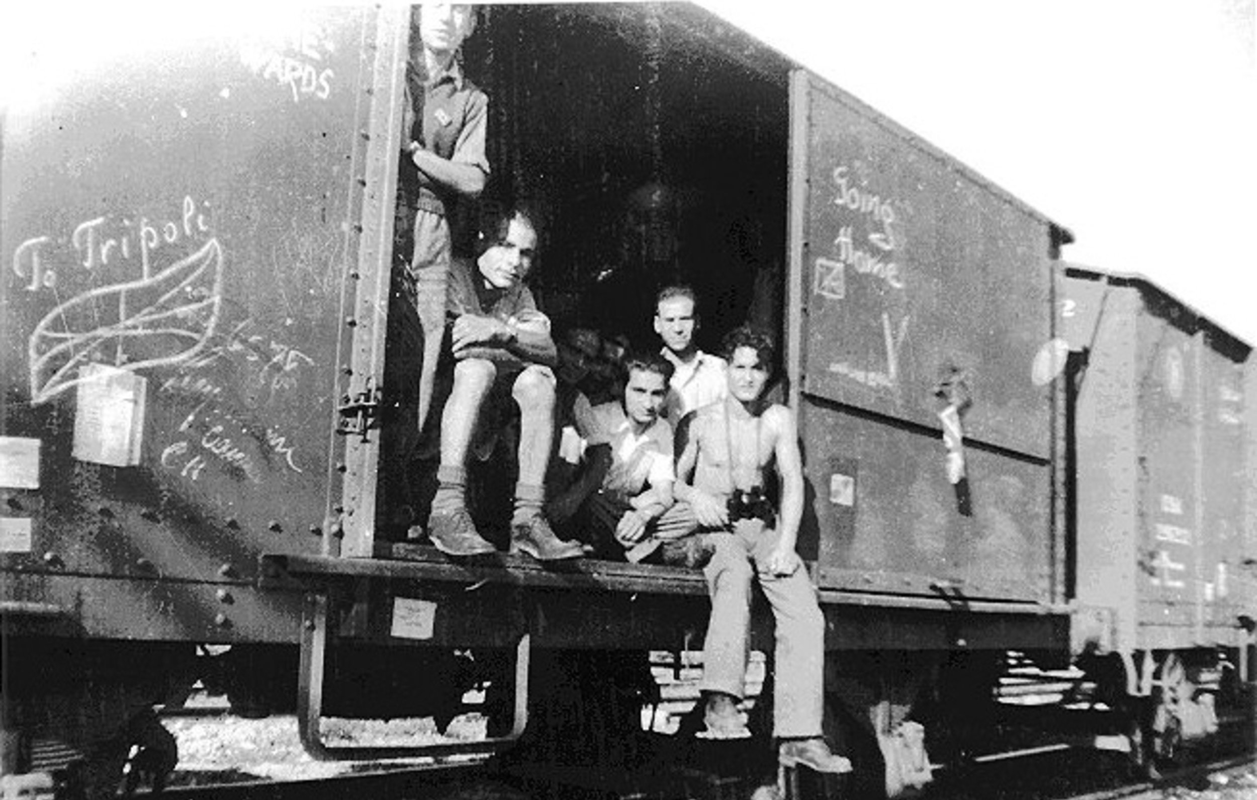 L'image montre un groupe de jeunes hommes assis et debout dans un wagon de train. Ils semblent se trouver dans un environnement rural ou industriel, avec des rails visibles au premier plan. Les murs du wagon sont couverts de graffitis, et l'ambiance générale paraît nostalgique. Les expressions des jeunes varient, allant de la réflexion à l'insouciance. L'image est en noir et blanc, ce qui lui donne un aspect vintage.
