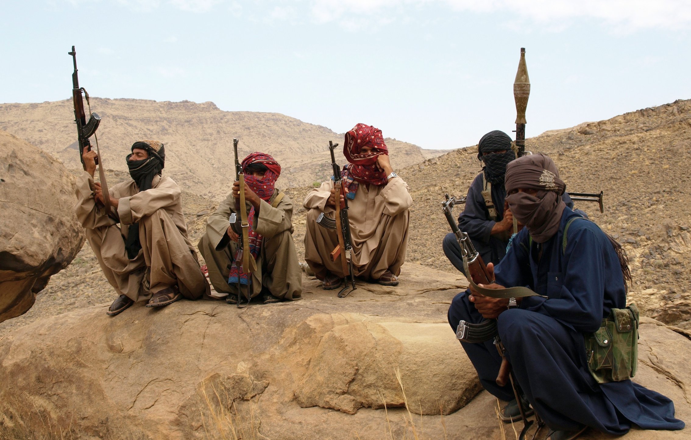 L'image montre un groupe de personnes assises sur des rochers dans un paysage désertique. Elles portent des vêtements traditionnels, masquent partiellement leur visage avec des foulards et tiennent des armes. L'environnement est montagneux et aride, avec des formations rocheuses en arrière-plan. L'ambiance semble sérieuse et reflète une situation de tension.