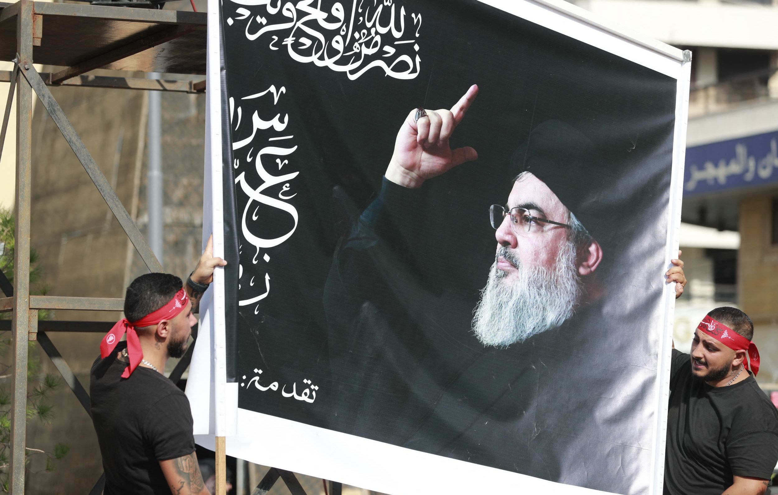 L'image montre deux personnes tenant une grande affiche. Cette affiche représente un homme avec une barbe, portant un vêtement sombre, et levant un doigt. En arrière-plan, on aperçoit des bâtiments. Les deux personnes portent des bandanas rouges et des t-shirts noirs. L'affiche comporte également des inscriptions arabes.