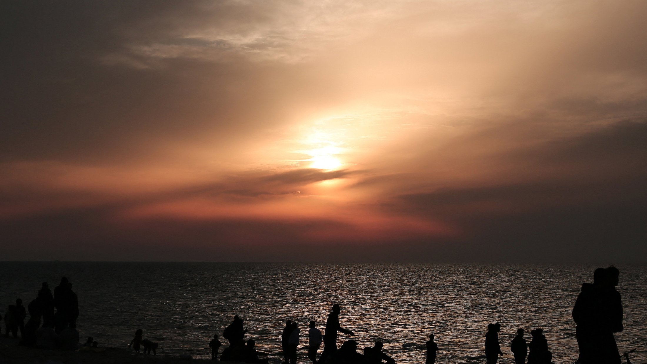 L'image montre une plage au crépuscule, avec des nuages qui tamisent la lumière du soleil couchant. De nombreuses silhouettes de personnes se déplacent le long du rivage, profitant de la scène. La mer reflète les couleurs douces du ciel, créant une atmosphère tranquille et paisible. Les silhouettes des individus ajoutent une dynamique à l'image, suggérant une ambiance conviviale et joyeuse.