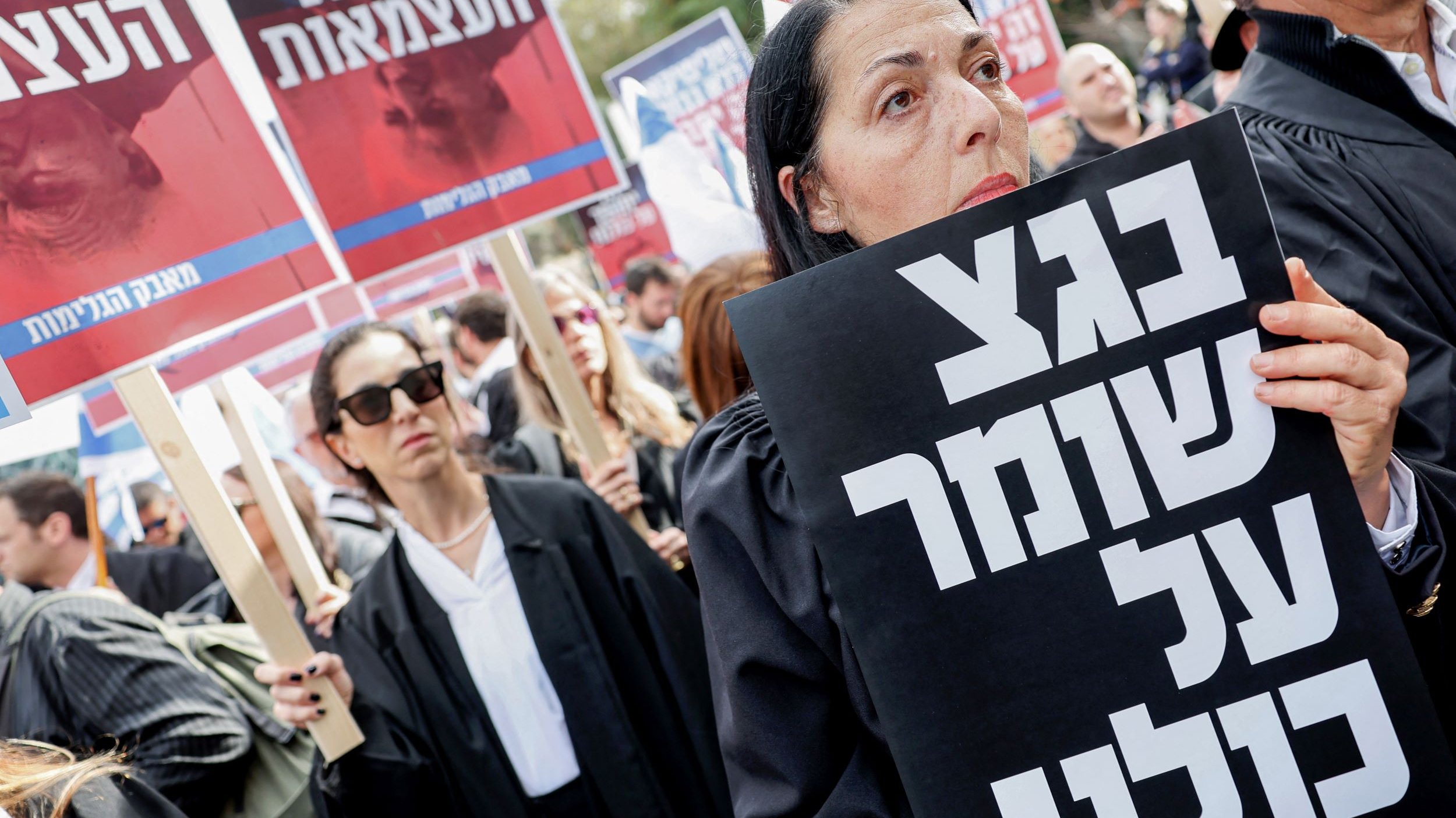 L'image montre une manifestation avec des participants portant des vêtements sombres, probablement des avocats, et tenant des pancartes. L'un des manifestants, au premier plan, tient une grande affiche noire avec du texte écrit en hébreu. L'atmosphère semble être sérieuse et engagée, avec des expressions résolues sur les visages des manifestants. En arrière-plan, d'autres personnes tiennent également des pancartes, ajoutant à l'ambiance de protestation.