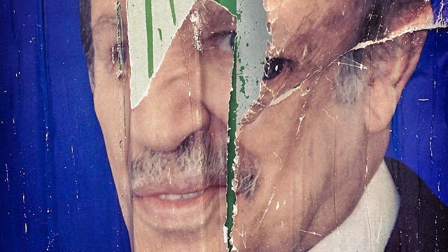 L'image montre un affichage déchiré d'un visage, probablement d'une personne publique, sur un fond bleu. Le visage est partiellement caché par des déchirures et des marques de pluie ou de peinture verte, ce qui donne un aspect usé et dégradé à l'image. Les détails du visage, comme les yeux et la bouche, sont visibles en partie, mais l'ensemble est affecté par le temps et l'usure.