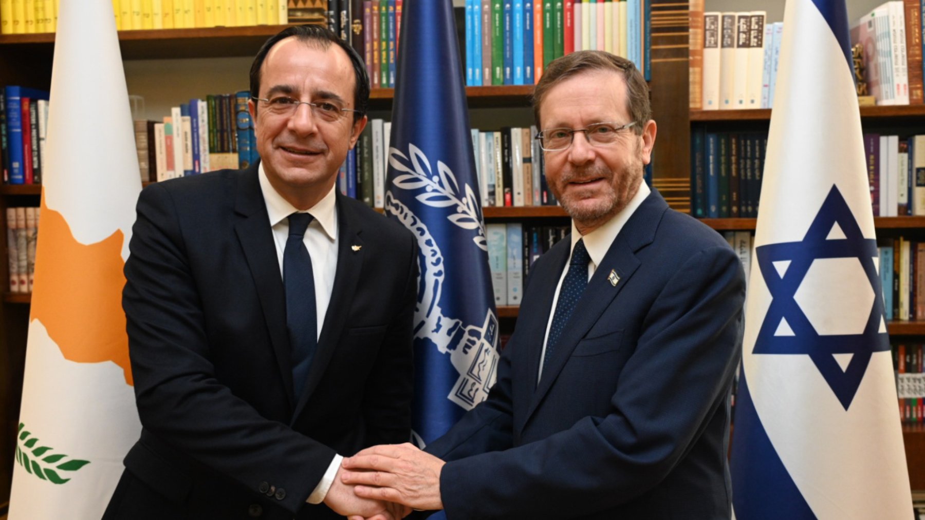Deux hommes en costume se serrent la main devant des drapeaux et des étagères de livres.