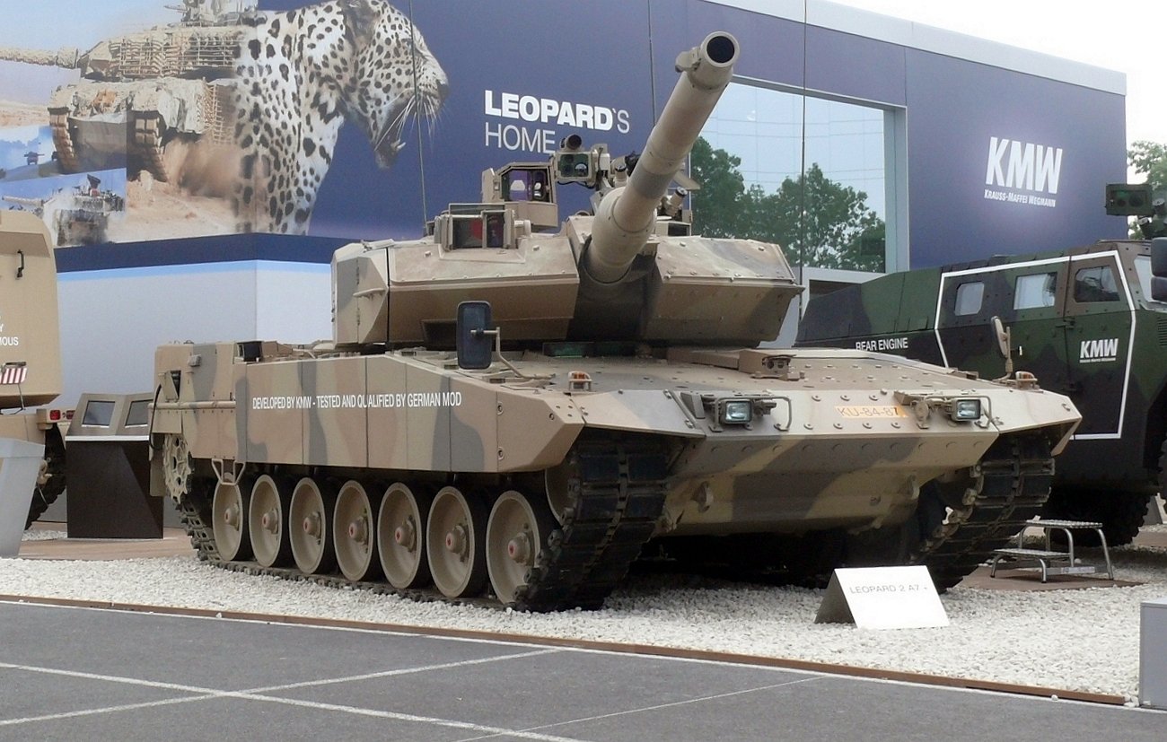 L'image montre un véhicule militaire, plus précisément un char de combat. Le char est peint dans un motif de camouflage beige et brun, typique pour se fondre dans des environnements désertiques. Il est équipé d'un canon long monté sur le dessus et possède plusieurs roues de chaque côté. En arrière-plan, on aperçoit des éléments d'exposition militaire, suggérant que le char est présenté lors d'un salon ou d'une exposition lié à la défense. Les détails tels que les plaques d'identification du char peuvent également être visibles.