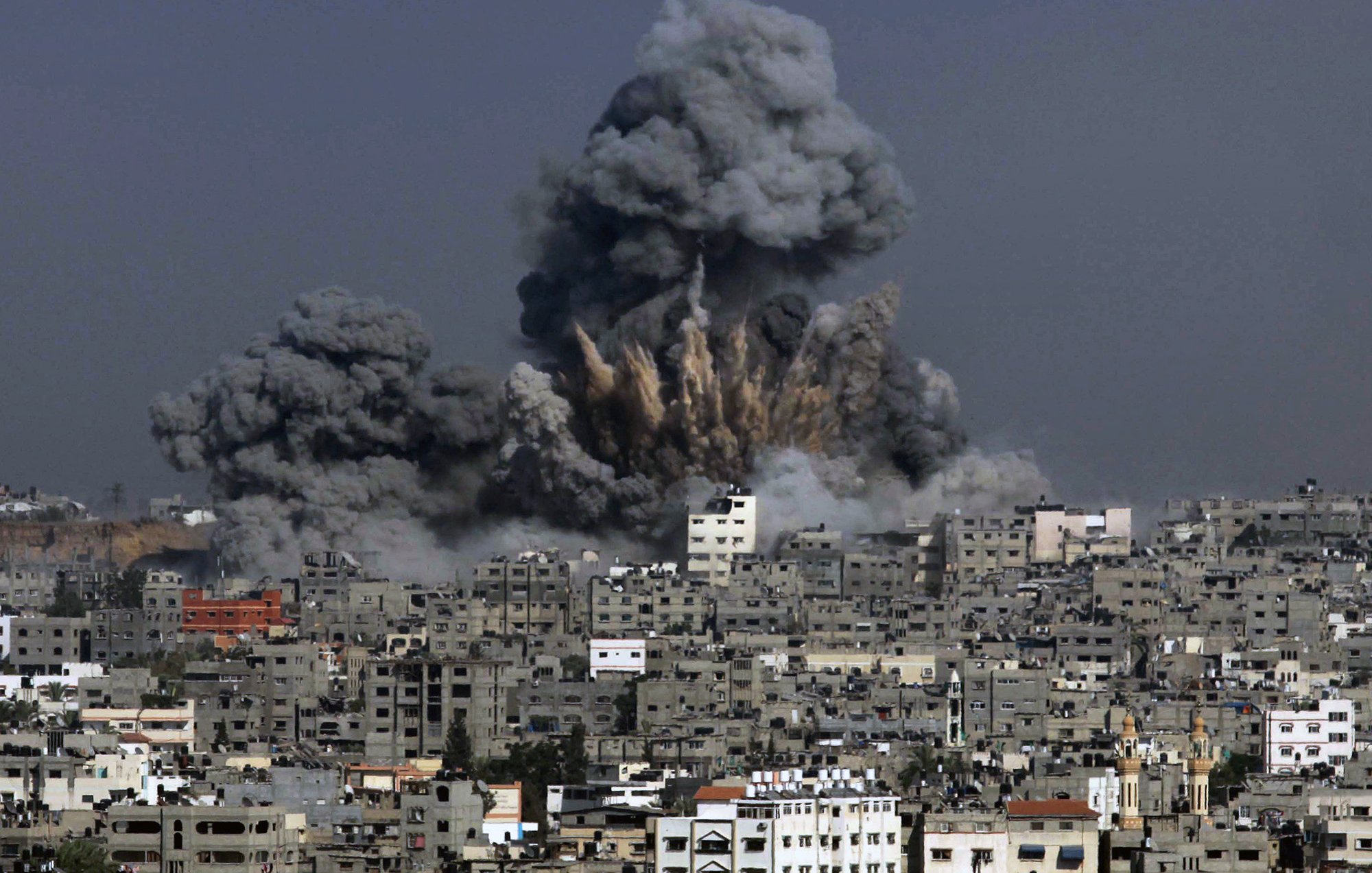 L'image montre une scène de destruction où des colonnes de fumée noire s'élèvent dans le ciel, indiquant qu'une explosion a eu lieu. Les bâtiments sont présents au premier plan, témoignant d'une zone urbaine, et semblent avoir subi des dommages. Le contraste entre la couleur grise de la fumée et les structures environnantes évoque une atmosphère de chaos et de conflit.