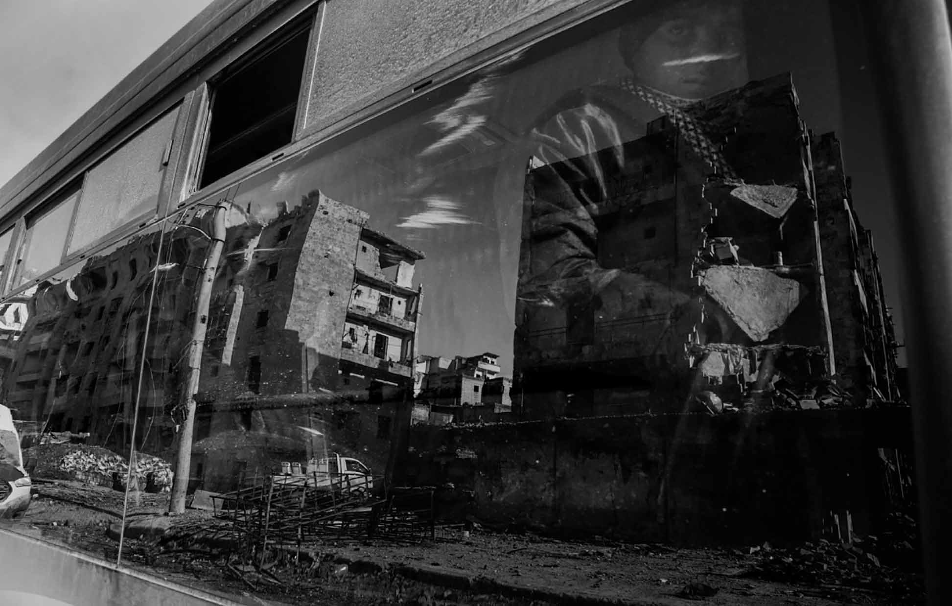 L'image présente une scène en noir et blanc, où un bus est au premier plan. On peut voir le reflet d'un homme, probablement en train de porter un vêtement sombre et une écharpe, à côté d'une architecture en ruine. À travers la vitre du bus, des bâtiments dégradés sont visibles, indiquant des signes de destruction. L'atmosphère semble sombre et tragique, évoquant des thèmes de dévastation et de résilience.