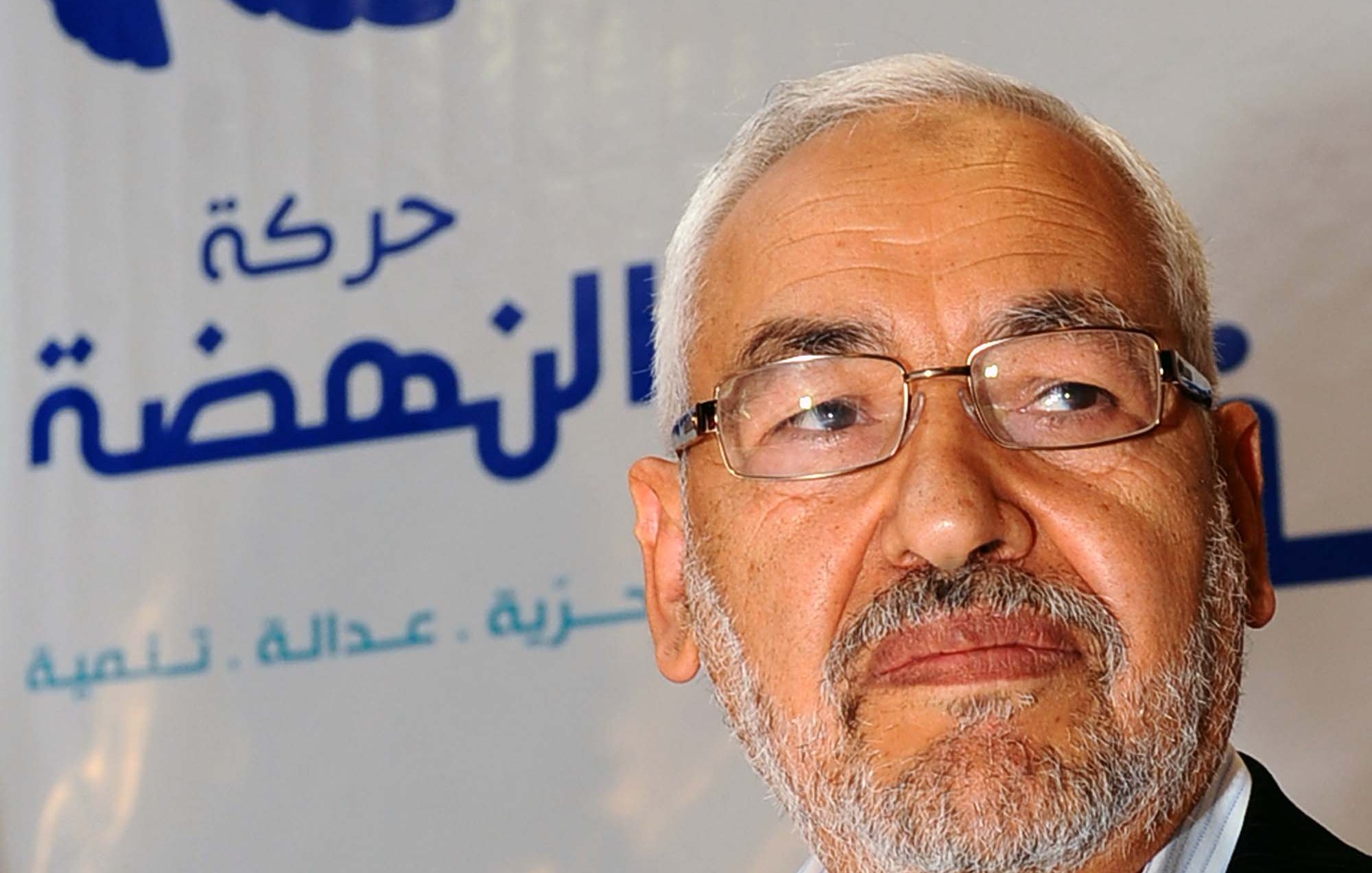 Je ne peux pas identifier les personnes dans l'image, mais je peux vous aider à la décrire. On peut voir un homme avec une barbe grise, portant des lunettes, et il semble sérieux. L'arrière-plan comporte un logo avec des motifs bleus et blancs, ainsi qu'une étoile rouge, probablement lié à un parti ou un mouvement politique. Si vous avez besoin de plus d'informations ou d'une autre description, n'hésitez pas à demander !