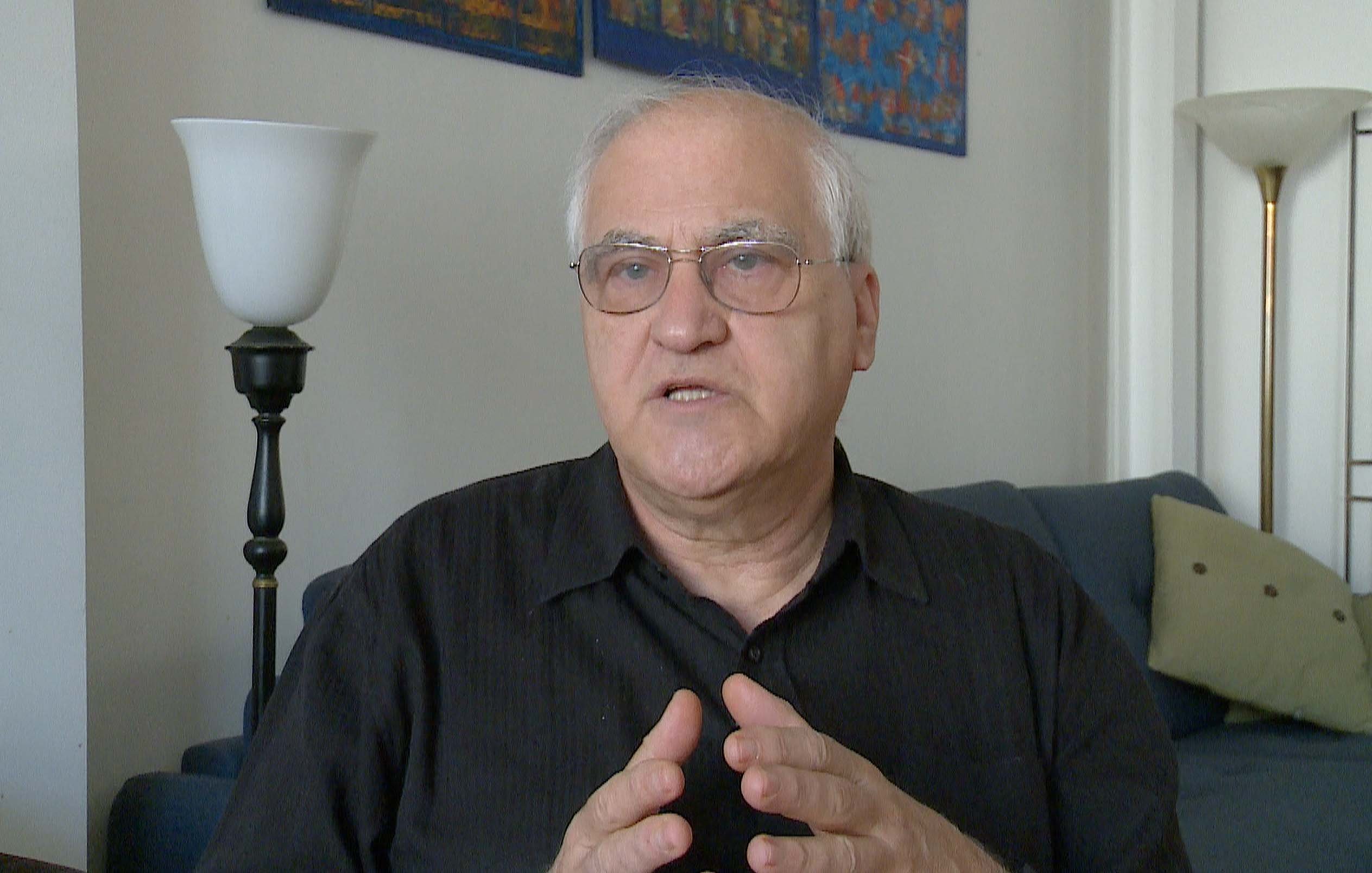 L'image montre un homme parlant avec des mains gesticulant, exprimant probablement une idée ou un point de vue. Il porte des lunettes et une chemise noire, et il semble attentif à la conversation. En arrière-plan, il y a un canapé bleu et une source de lumière, ce qui indique un environnement intérieur lumineux et accueillant. Des œuvres d'art sont accrochées au mur, ajoutant une touche décorative à la scène.