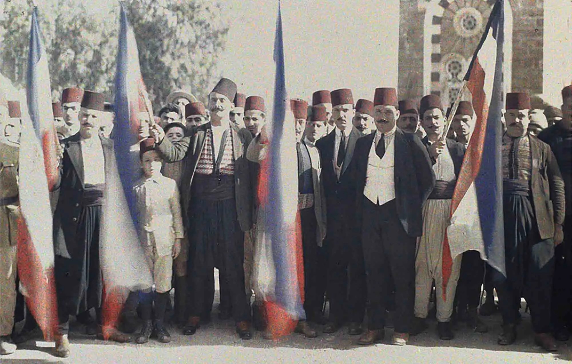 L'image montre un groupe de personnes en tenue traditionnelle, se tenant ensemble avec des drapeaux russes. Les hommes portent des fez et divers vêtements folkloriques, avec un jeune garçon au centre. L'arrière-plan présente une architecture qui semble historique, peut-être un bâtiment symbolique. L'atmosphère suggère un moment de célébration ou de rassemblement associé à une certaine fierté nationale.