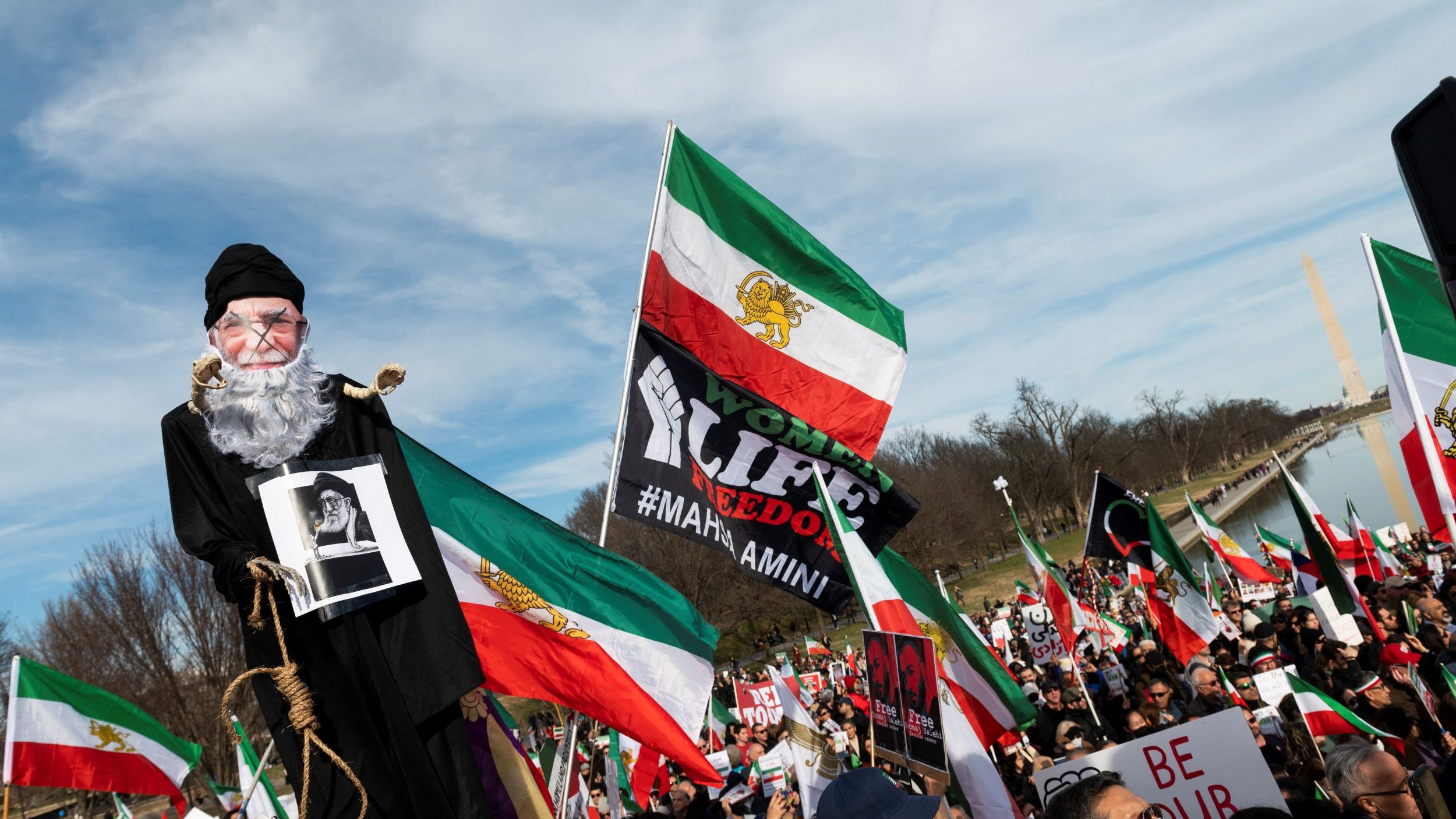 L'image montre une grande manifestation, probablement en faveur de la liberté ou des droits de l'homme. Au premier plan, une marionnette représentant un homme âgé est portée par un manifestant, tandis que d'autres personnes tiennent des drapeaux iraniens et des pancartes. Les participants semblent unis et déterminés, exprimant leurs revendications. L'ambiance est dynamique, avec une foule nombreuse et des symboles visuels forts liés à leur cause.