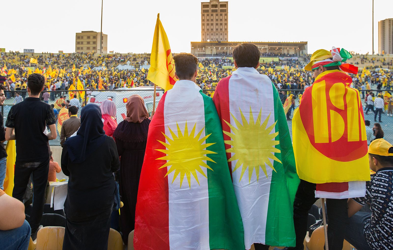 L'image montre un événement festif dans un stade rempli de supporters. Au premier plan, deux personnes portent des drapeaux, l'un étant le drapeau kurde avec les étoiles et l'autre avec un symbole qui semble représenter un mouvement ou un parti. Le public derrière eux est également vêtu de couleurs vives, principalement du jaune, suggérant une ambiance célébratoire. La scène évoque un fort sentiment d'unité et de fierté culturelle.