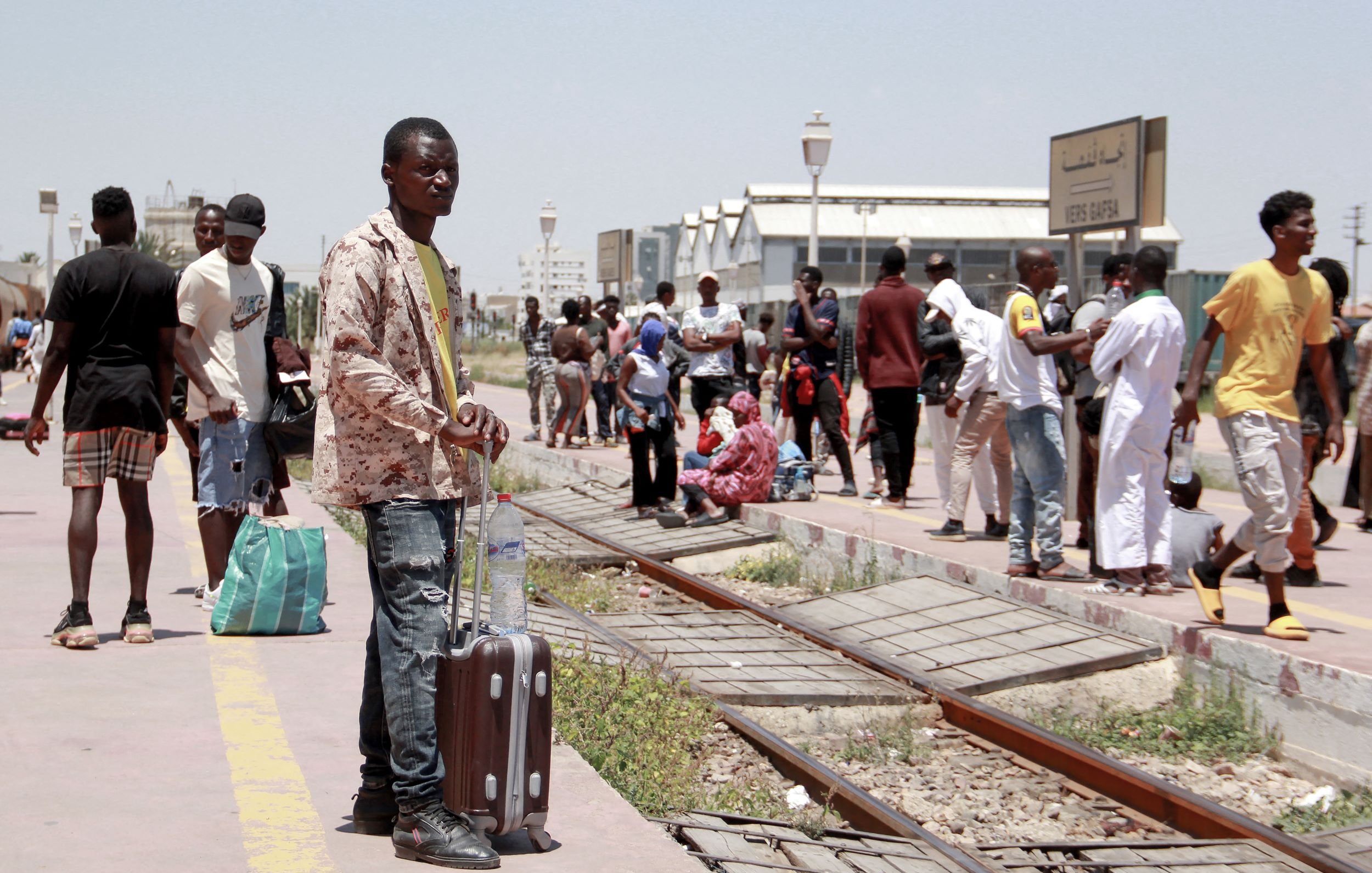 L'image montre une station de train animée. Un homme debout, tenant une valise, semble attendre. En arrière-plan, on peut voir d'autres personnes : certaines attendent assises, d'autres se déplacent avec des sacs. L'environnement est ensoleillé, et il y a des bâtiments visibles au loin. La scène évoque un moment de déplacement et d'attente.