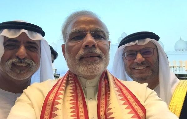 L'image montre un groupe de trois hommes prenant un selfie, probablement dans un cadre officiel ou lors d'un événement. L'homme au centre porte un vêtement traditionnel avec un foulard coloré autour du cou. Les deux autres hommes à côté de lui portent des vêtements traditionnels du Moyen-Orient, incluant un keffieh et un costume, ajoutant à l'atmosphère culturelle de la scène. En arrière-plan, on aperçoit des bâtiments qui semblent être emblématiques, bien que les détails ne soient pas clairs. L'expression des visages est souriante et conviviale, suggérant une atmosphère amicale.