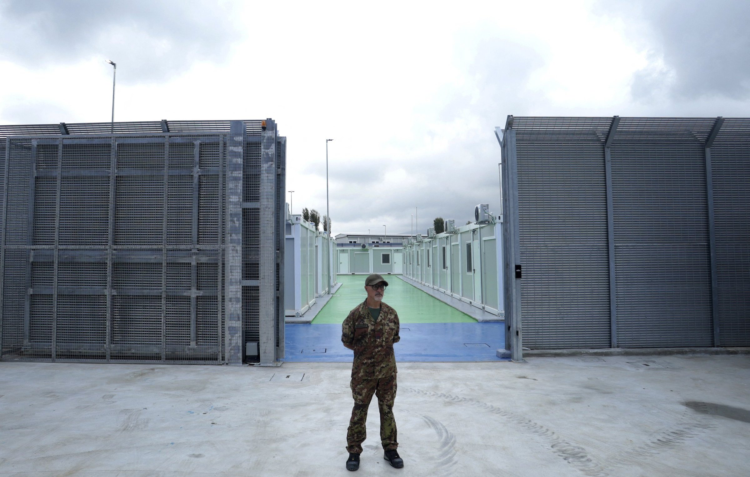 L'image montre un soldat en uniforme qui se tient au centre d'une zone sécurisée, probablement à l'extérieur d'une installation. À l'arrière-plan, on peut voir des structures métalliques grises et des murs, créant une ambiance de confinement. Le sol est en béton, et il y a une passerelle colorée qui semble être verte ou bleue. Le ciel est nuageux, suggérant un temps sombre. L'ensemble de l'image évoque un sentiment de sécurité renforcée et de surveillance.