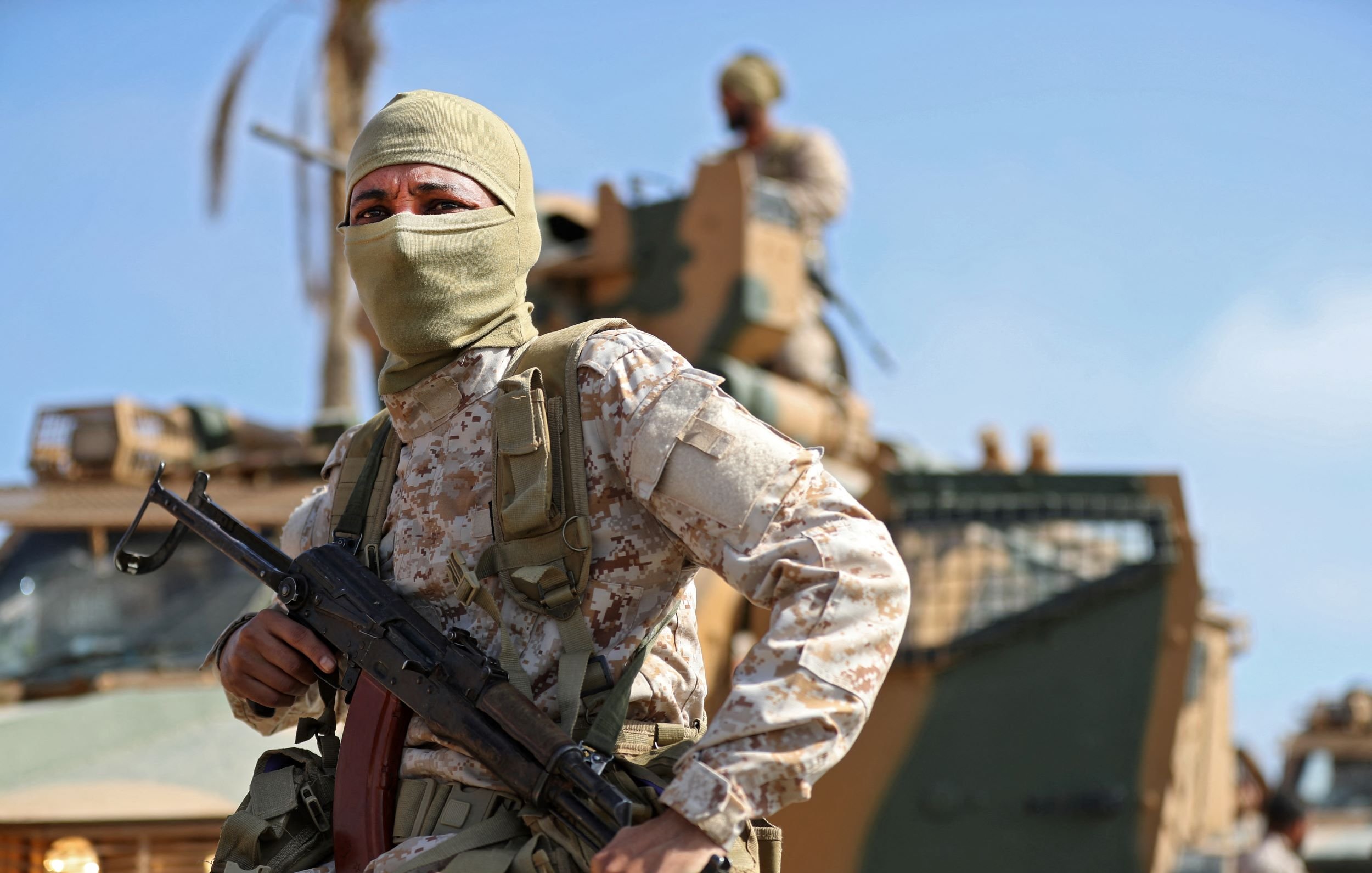 L'image montre un soldat portant un uniforme camouflé et un masque couvrant le bas de son visage. Il est équipé d'une arme et se tient en position vigilante, probablement en mission. En arrière-plan, on peut apercevoir un véhicule militaire et un autre soldat. Le ciel est dégagé, suggérant une ambiance ensoleillée et une atmosphère de tension.