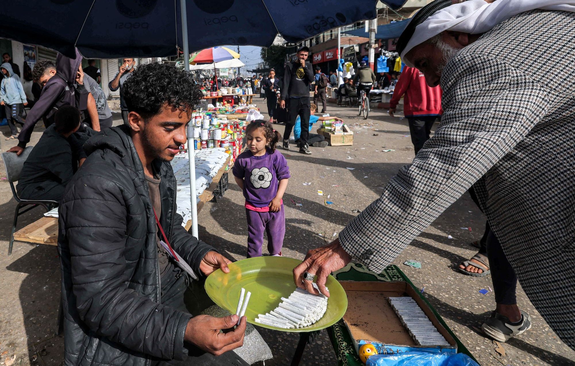 L'image montre une scène de marché dans une rue animée. Un jeune homme est assis sur un banc, tenant une assiette ronde verte sur ses genoux, où semblent être disposés des articles, probablement des bonbons ou des bâtonnets. Un homme plus âgé s'approche de lui, tenant quelque chose dans sa main, peut-être en train de lui faire une offre ou de lui donner des articles. En arrière-plan, il y a des gens qui se déplacent, ainsi que des étals avec divers produits. Une petite fille se tient à proximité, observant la scène. Les parasols créent une ambiance de marché, tandis que l'atmosphère semble vivante et animée.