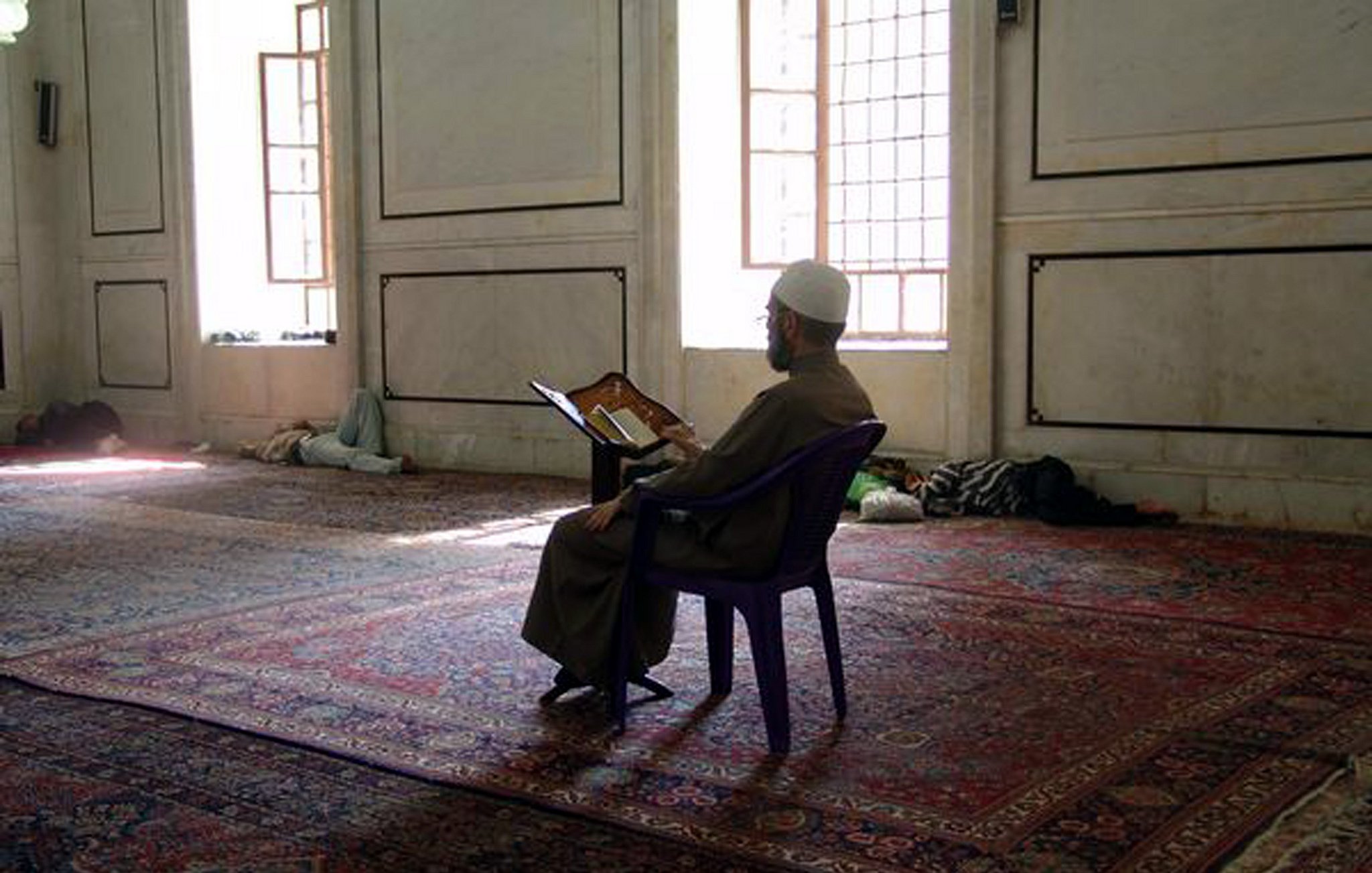 L'image montre un homme assis sur une chaise dans une mosquée. Il est vêtu d'une longue robe et porte un chapeau traditionnel. Devant lui, il lit un livre ouvert, probablement un exemplaire du Coran. La mosquée a un intérieur simple, avec des murs en marbre et des fenêtres laissant passer la lumière naturelle. Le sol est recouvert de tapis, et on peut voir d'autres personnes allongées au fond de la salle.
