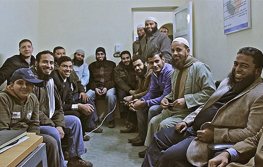 L'image montre un groupe d'hommes assis dans une pièce, probablement un bureau ou un espace de réunion. Ils semblent être de différentes origines et portent des vêtements variés, certains avec des chapeaux ou des turbans. Les participants affichent des expressions amicales et sont rassemblés de manière à discuter ou à partager un moment ensemble. L'ambiance semble conviviale et détendue.