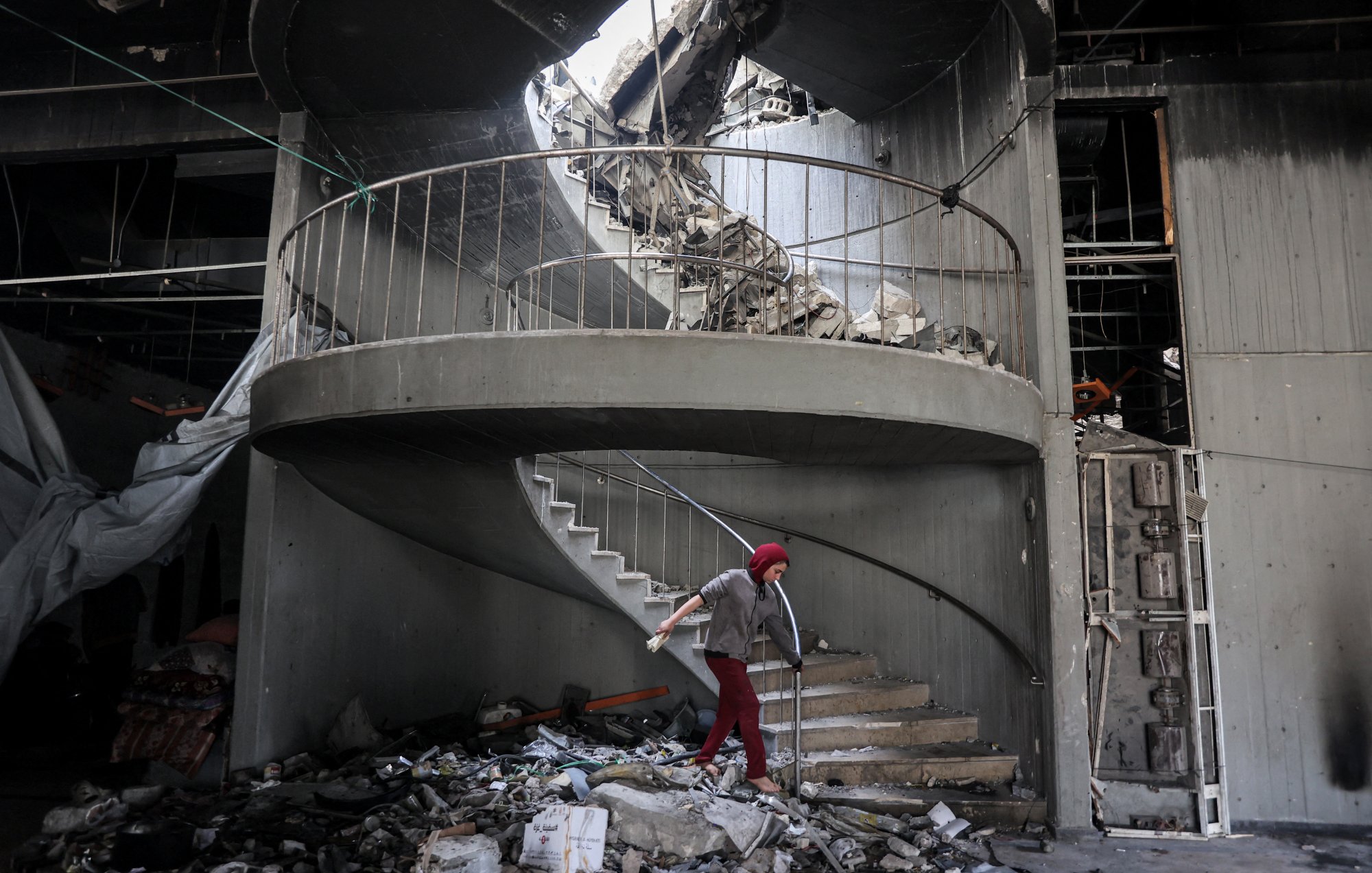 L'image montre un escalier en colimaçon partiellement détruit, avec des débris éparpillés autour. Le plafond a subi d'importants dégâts, laissant apparaître un trou par lequel la lumière pénètre. Un individu, utilisant des béquilles, se déplace prudemment sur les marches. L'ambiance générale évoque des conditions de délabrement et de désolation, probablement à la suite d'un événement catastrophique.