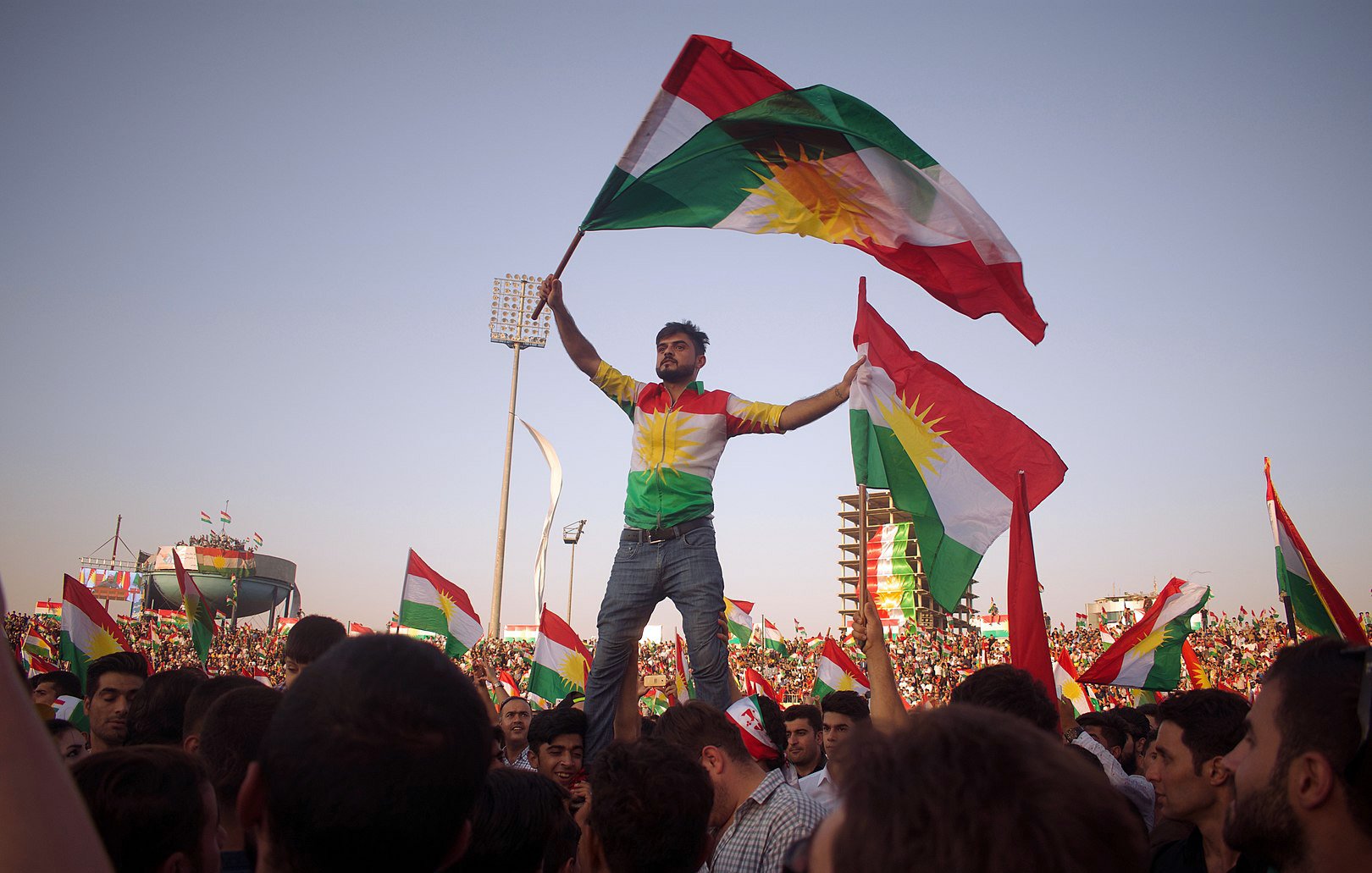 L'image montre une grande foule célébrant, probablement lors d'un événement culturel ou politique. Au premier plan, un homme se tient debout sur une autre personne, tenant des drapeaux kurdes. Il porte un maillot aux couleurs du drapeau kurde, et son expression semble joyeuse et enthousiaste. En arrière-plan, on peut voir de nombreuses autres personnes agitant également des drapeaux, créant une atmosphère festive et énergétique. Le ciel est clair, ce qui ajoute à l'ambiance dégagée de la scène.