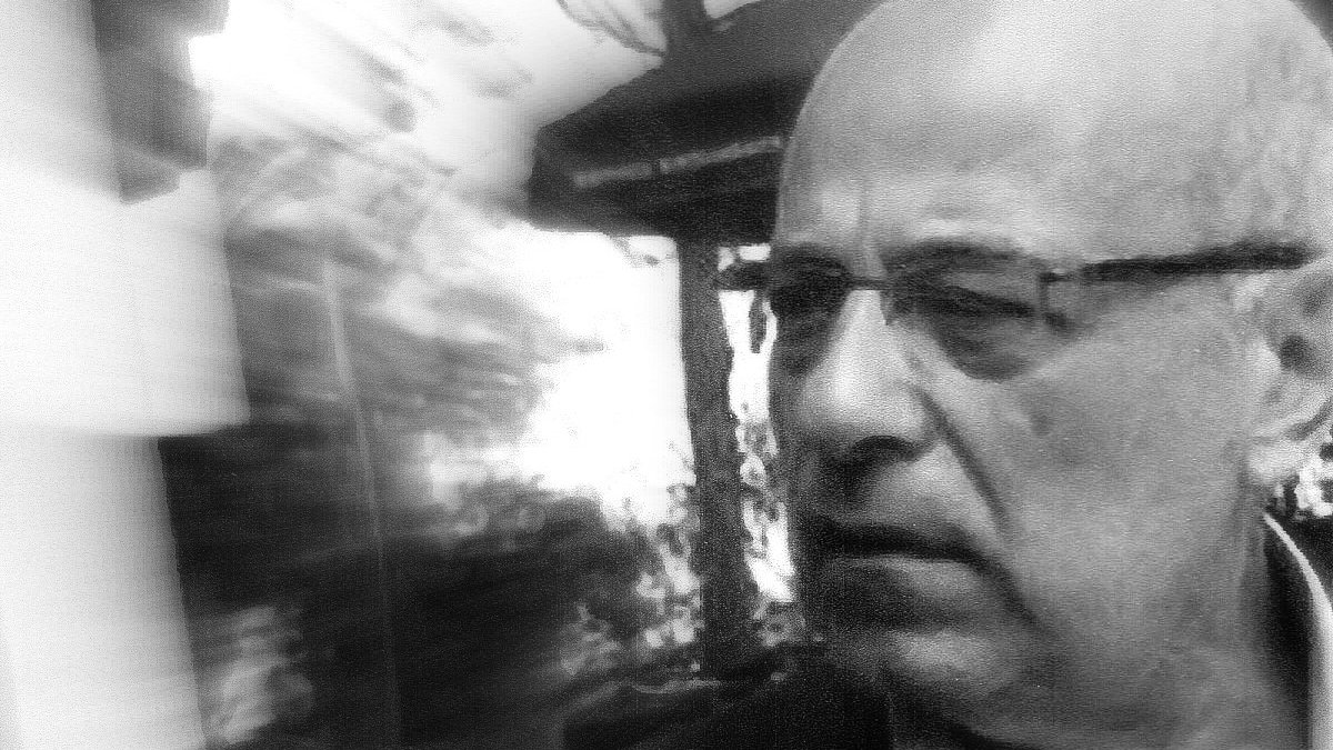 L'image présente un homme d'âge mûr avec des lunettes, légèrement flou en raison d'un effet de mouvement. Il semble pensif ou préoccupé, et son expression faciale transmet une certaine intensité. L'arrière-plan est indistinct, ce qui ajoute à l'effet dynamique de la photo. Les tonalités en noir et blanc renforcent le caractère dramatique de l'image.