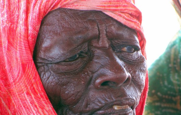 L'image montre un portrait d'une personne âgée avec une expression réfléchie. Elle porte un foulard coloré, qui semble être enroulé autour de sa tête, et son visage présente des rides marquées, témoignant de son âge et de son vécu. La lumière met en valeur les détails de son visage.