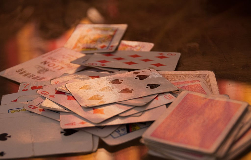 L'image montre un tas de cartes à jouer éparpillées sur une surface. On peut voir différents types de cartes, y compris des cartes de cœur, de carreau, de trèfle et de pique. Certaines cartes semblent retournées, tandis que d'autres sont face visible, montrant leurs motifs et valeurs. L'éclairage apporte une ambiance chaleureuse et met en valeur la texture des cartes.