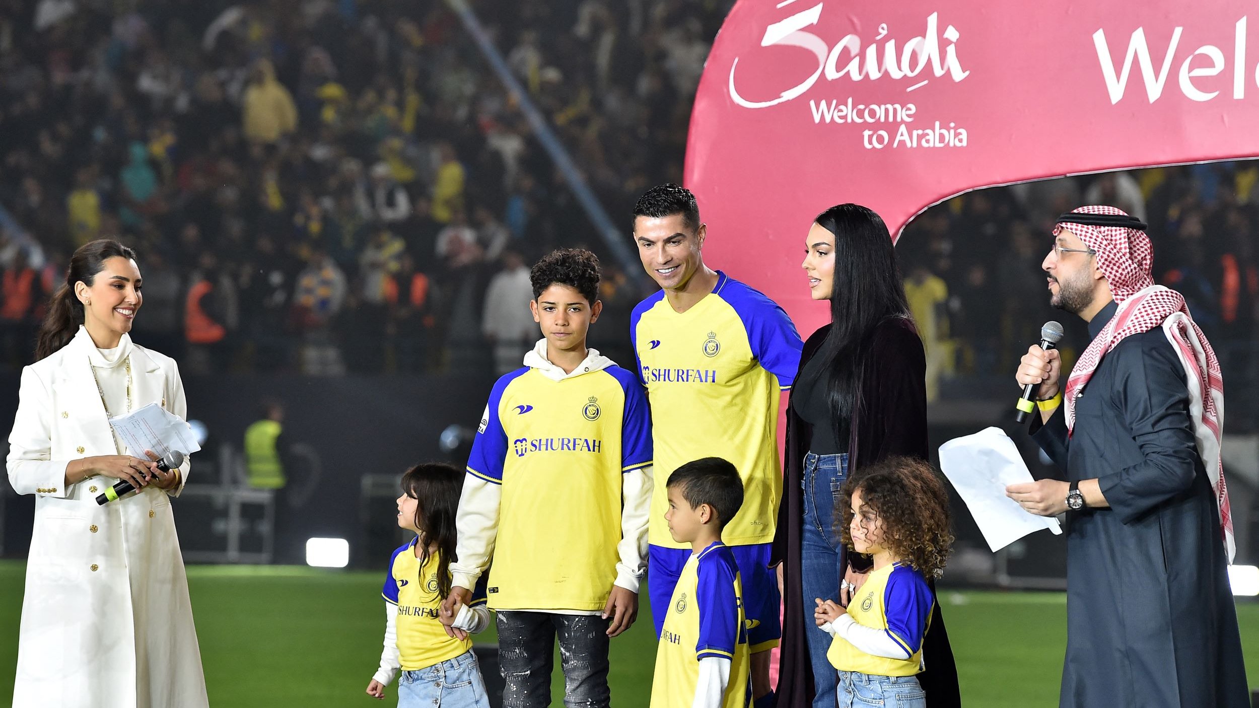 L'image montre une scène sur un terrain de football, où plusieurs personnes sont réunies autour d'un joueur, probablement un footballeur célèbre, en tenue de jeu. Il y a des enfants, probablement des membres de la famille, qui se tiennent à ses côtés. Une femme, habillée d'une manière élégante, semble s'adresser à l'audience, tandis qu'un homme en tenue traditionnelle parle également en tenant un microphone. En arrière-plan, des spectateurs sont visibles, et une grande arche avec les mots "Welcome to Arabia" est installée, indiquant un événement spécial.