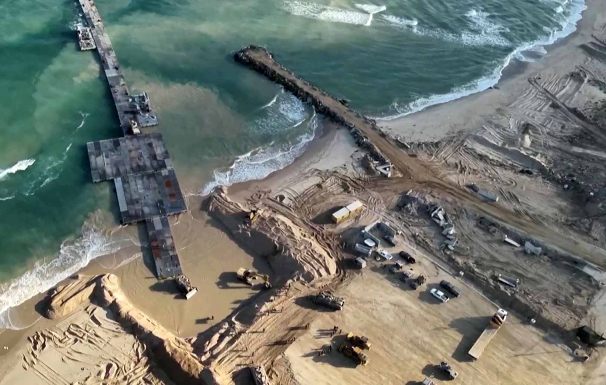 L'image montre une vue aérienne d'un site de construction situé près de la mer. On peut voir un port ou une jetée inachevée s'étendant dans l'eau, entouré de terres travaillées. Des engins de construction sont dispersés autour, et le sol est marqué par des travaux de terrassement. Les vagues de la mer s'échouent à proximité, créant un contraste avec la zone de construction. L'ensemble de la scène illustre une zone en développement, probablement pour une infrastructure côtière.