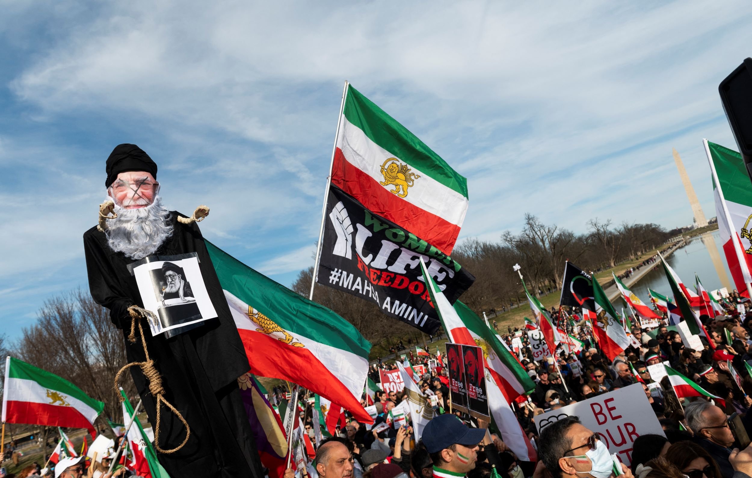 L'image montre une grande manifestation, probablement en faveur de la liberté ou des droits de l'homme. Au premier plan, une marionnette représentant un homme âgé est portée par un manifestant, tandis que d'autres personnes tiennent des drapeaux iraniens et des pancartes. Les participants semblent unis et déterminés, exprimant leurs revendications. L'ambiance est dynamique, avec une foule nombreuse et des symboles visuels forts liés à leur cause.