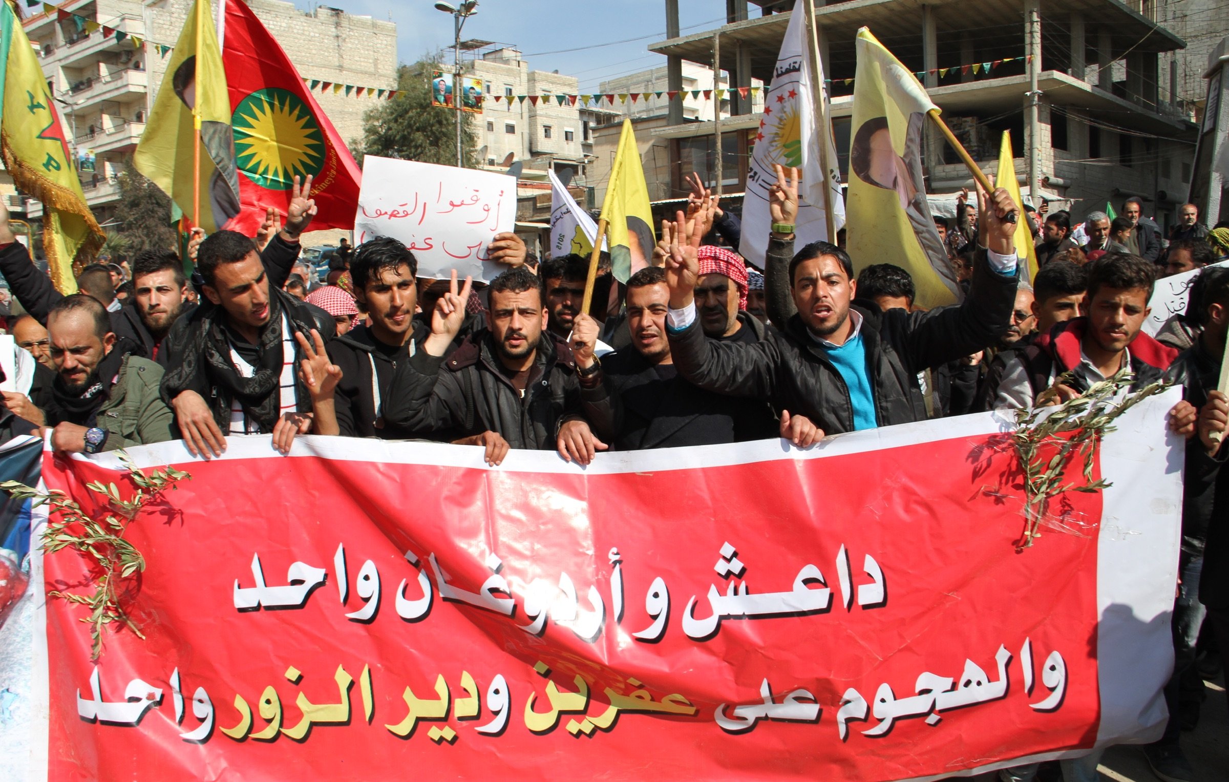 L'image montre un groupe de manifestants tenant des pancartes et des drapeaux, en train de défiler dans une rue. Ils portent des expressions déterminées et semblent militer pour une cause. Leurs vêtements et les drapeaux indiquent une provenance régionale. Une grande banderole rouge est affichée, avec des inscriptions en arabe. Les manifestants font le signe de la victoire avec leurs doigts, ce qui suggère un esprit de solidarité et de lutte. L'ambiance générale semble dynamique et engagée.