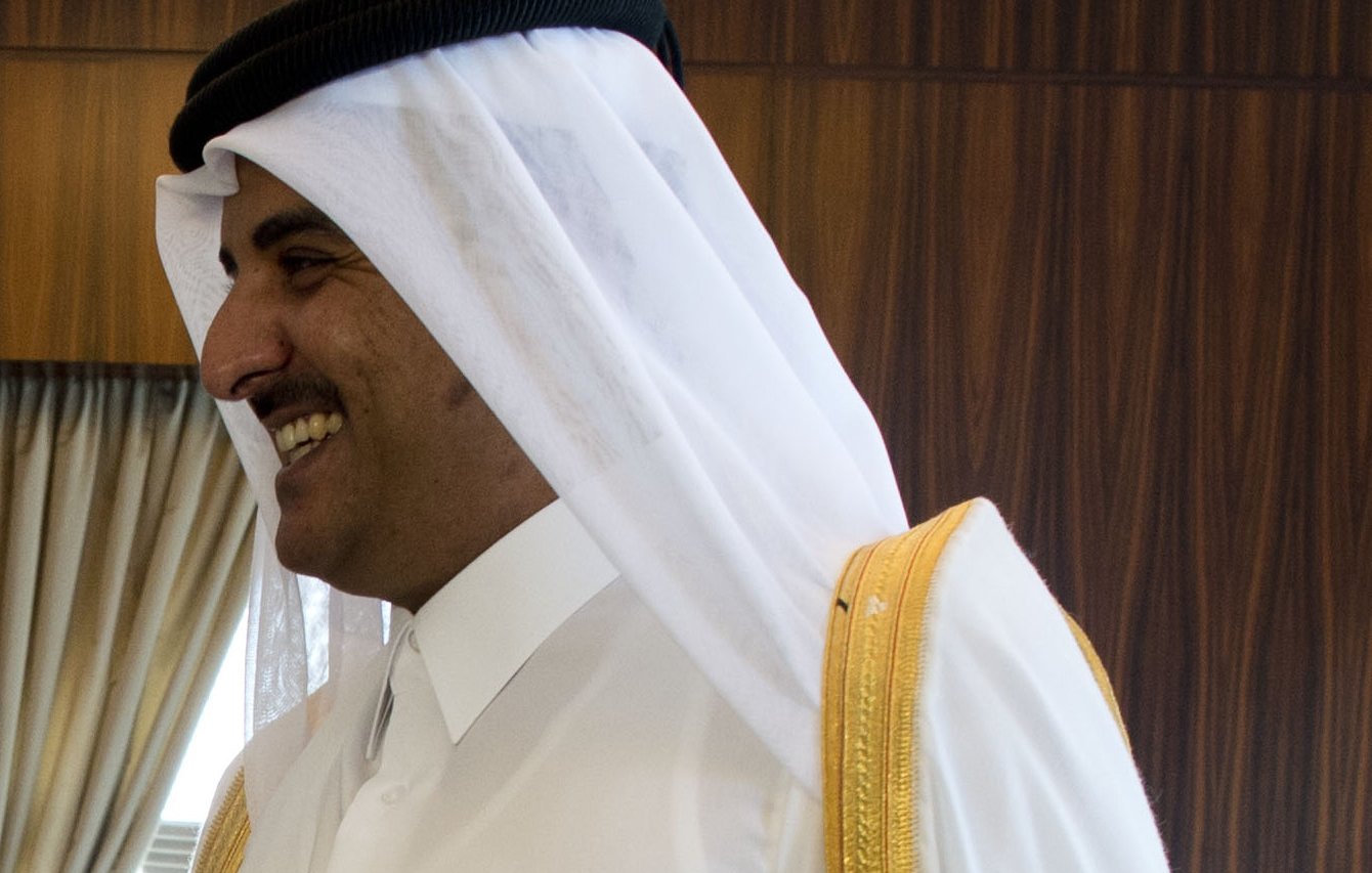 L'image montre un homme portant une tenue traditionnelle qatarienne. Il a un large sourire et semble être en mouvement, peut-être en train de parler ou d'interagir avec d'autres personnes. Sa tenue est composée d'une robe blanche, avec des ornements dorés, et il porte également un turban blanc avec un bandeau noir. L'arrière-plan montre des draperies et un environnement intérieur, indiquant un cadre formel ou officiel.