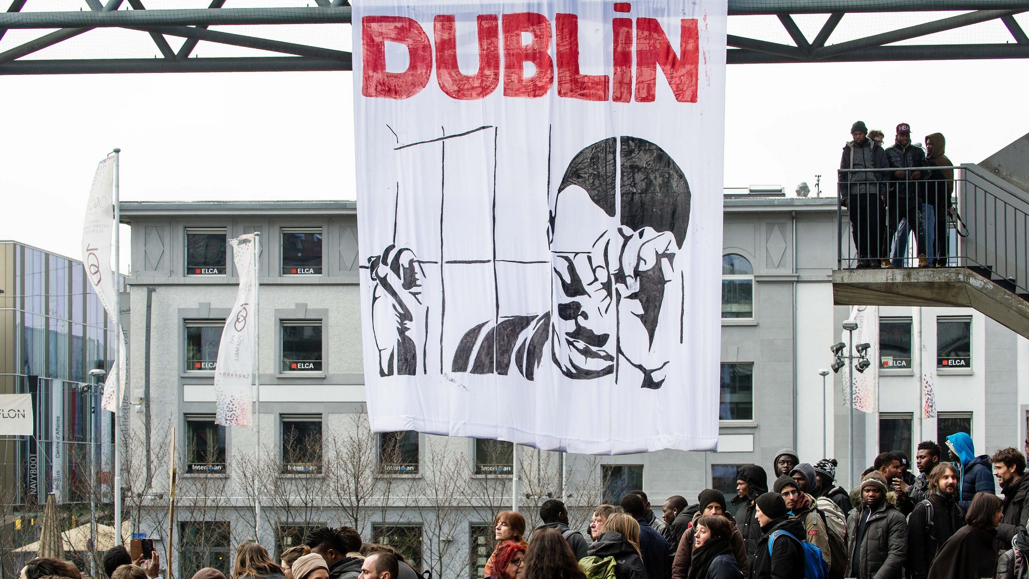 L'image montre une manifestation à Dublin, avec une grande banderole suspendue qui représente une figure tenant des barres comme une prison. En arrière-plan, on peut voir un groupe de personnes rassemblées, manifestant sur une route ou une place. Les gens semblent engagés dans la cause, et l'atmosphère est dynamique, probablement en réponse à une problématique sociale ou politique.