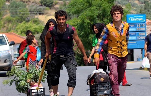 L'image montre un groupe de personnes se déplaçant le long d'une route. Il y a des enfants et des adultes, certains portent des vêtements colorés. Deux hommes avancent avec des paniers à roulettes, contenant probablement des biens ou de la nourriture. L'un d'eux tient une plante, tandis que l'autre semble porter une baguette ou un long objet. On peut voir des maisons et des arbres en arrière-plan, ajoutant une touche de nature à la scène. L'ambiance générale évoque un déplacement ou un voyage.