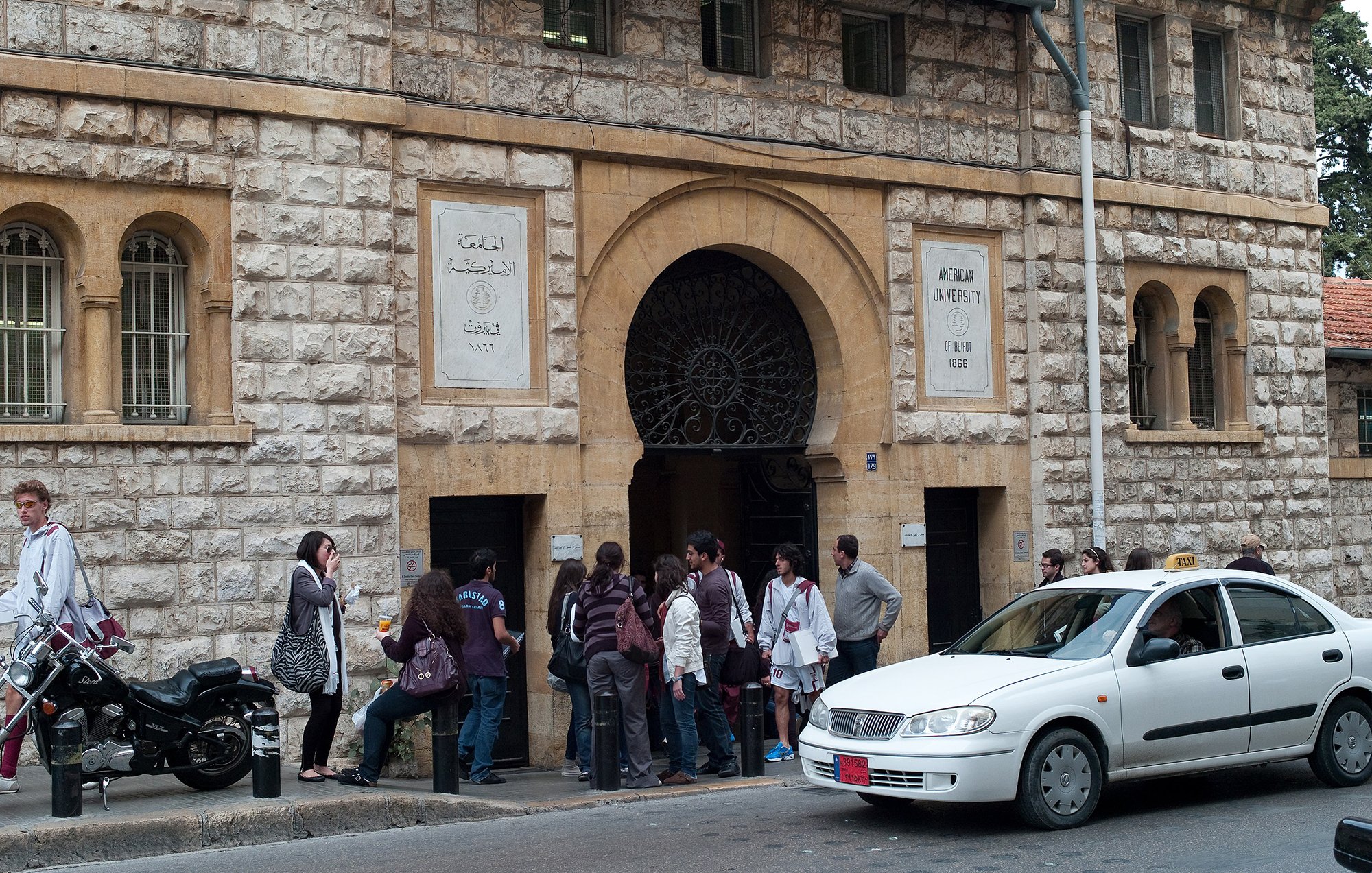 L'image montre l'entrée d'un bâtiment en pierre, probablement une université. Plusieurs personnes sont rassemblées devant l'entrée, créant une petite file d'attente. On peut voir des étudiants, certains portant des sacs. À droite, une voiture blanche passe proche de la scène, tandis qu'une moto est garée à proximité. L'architecture du bâtiment présente des éléments classiques avec un grand portail en fer.