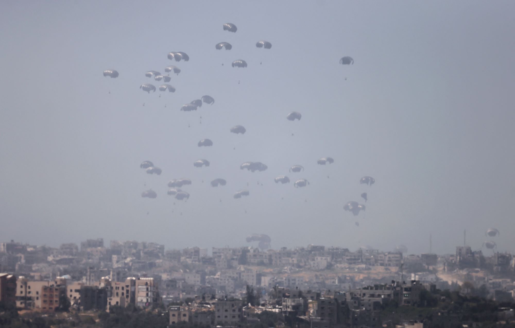 L'image montre un paysage urbain où de nombreux parachutes descendent lentement vers le sol. On peut voir une ville en arrière-plan, avec des bâtiments disposés de manière variable. L'atmosphère semble brumeuse, ce qui donne un aspect à la fois dramatique et intrigant à la scène. Les parachutes indiquent probablement une opération militaire ou humanitaire, créant un contraste avec l'environnement urbain.