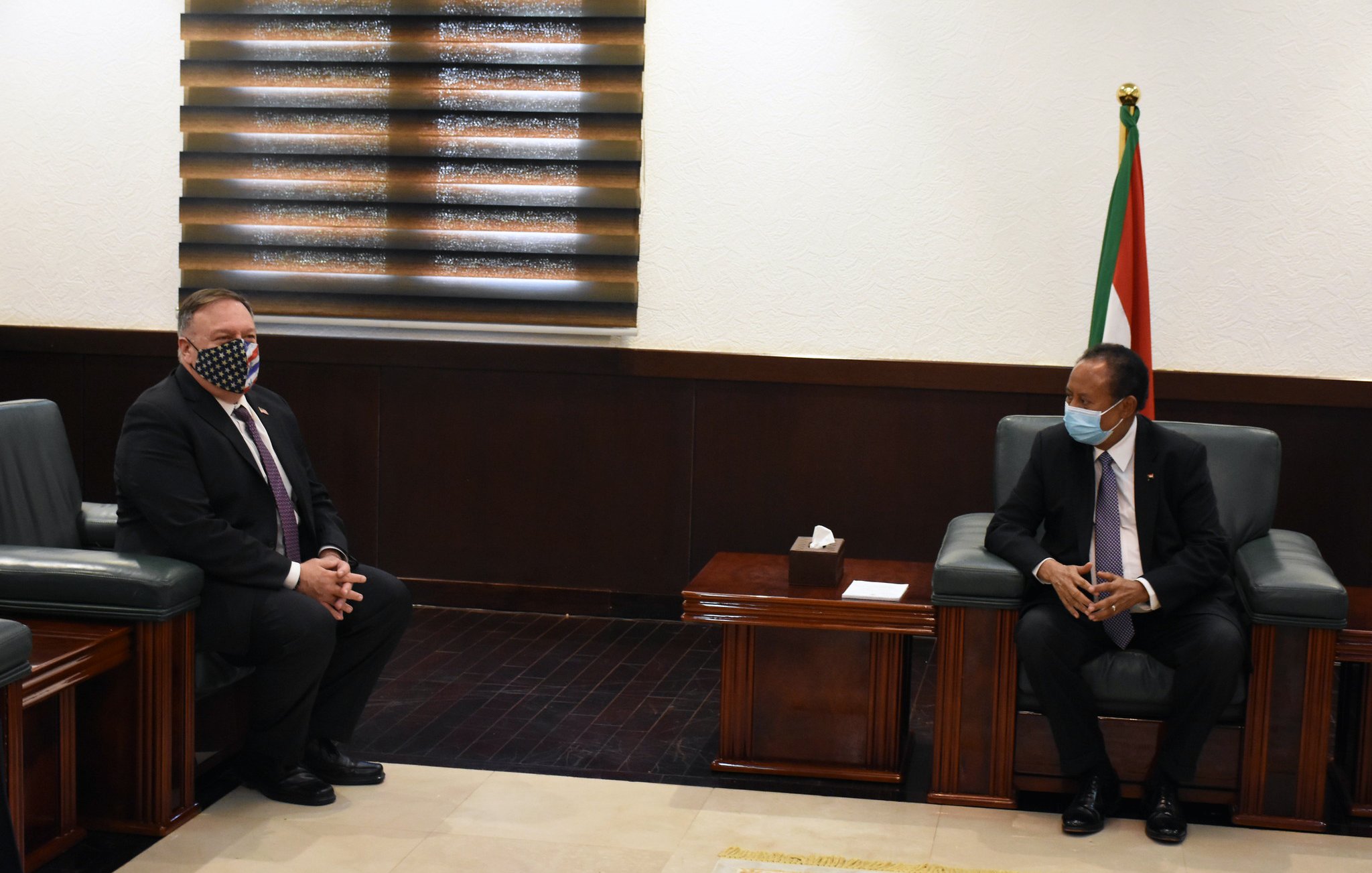 L'image montre deux hommes assis dans une pièce bien éclairée, avec des murs clairs et un sol en marbre. L'un des hommes porte un masque facial à motifs, tandis que l'autre est en costume avec une cravate. À l'arrière-plan, on peut voir un drapeau. Les deux hommes semblent engagés dans une conversation ou une réunion. Les meubles sont en bois sombre, et la pièce dégage une atmosphère formelle.