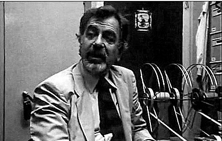 L'image présente un homme assis dans un bureau. Il porte un costume clair et une cravate foncée. Son expression faciale semble animée, presque passionnée, comme s'il était en train de discuter ou d'expliquer quelque chose. À l'arrière-plan, on peut voir des bobines de film ou des documents, ce qui donne une impression d'environnement de travail lié à la production ou à la recherche. L'éclairage est sombre, créant une ambiance vintage, typique des années passées.